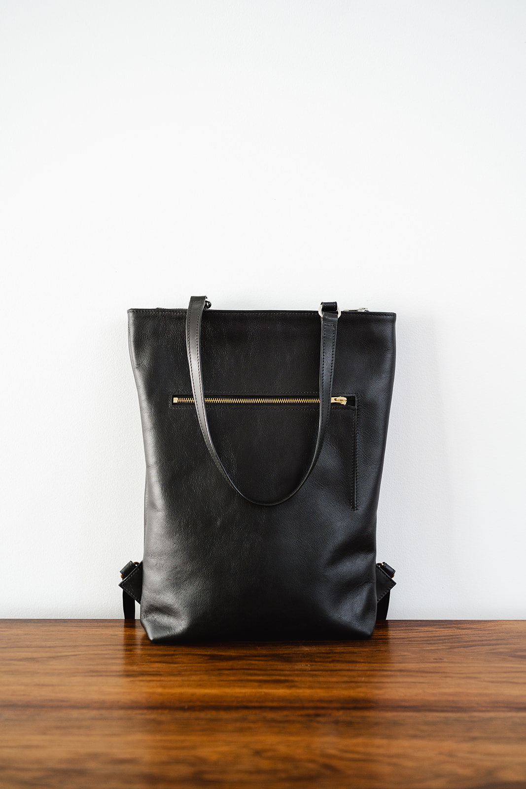 The Backpack by Ella Jackson Australia – minimalist black leather backpack standing upright against a white wall on timber table.