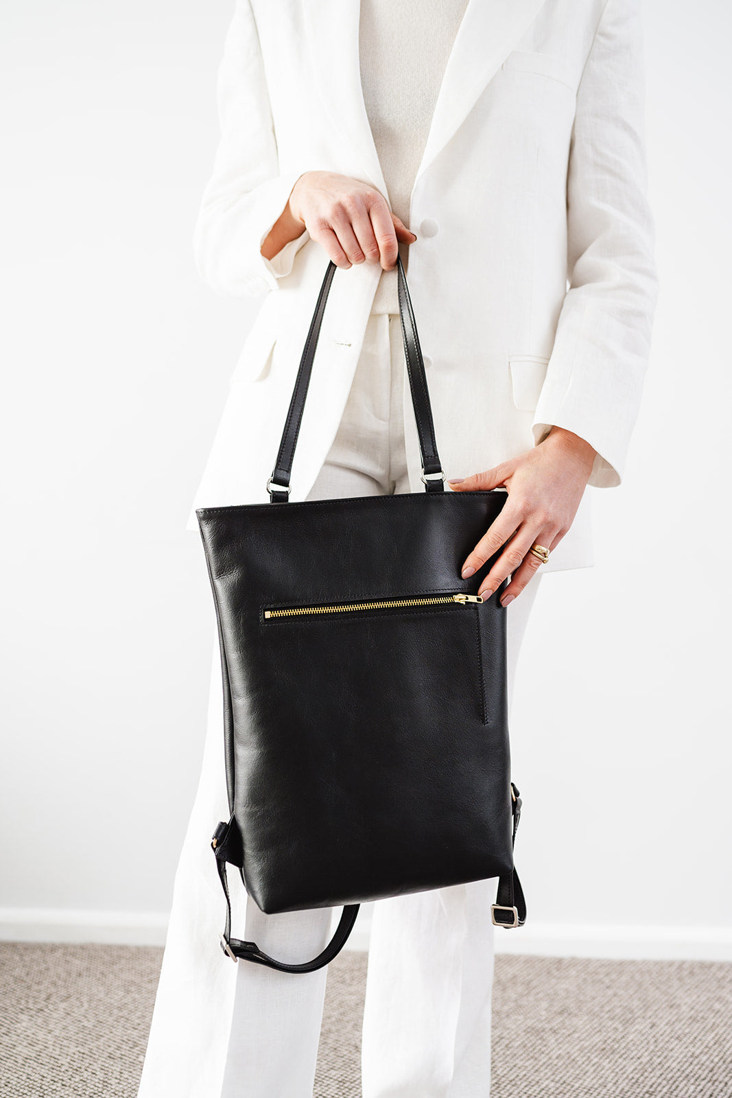 Close-up of The Backpack by Ella Jackson Australia – minimalist black leather backpack held by top handles, worn with a white suit.