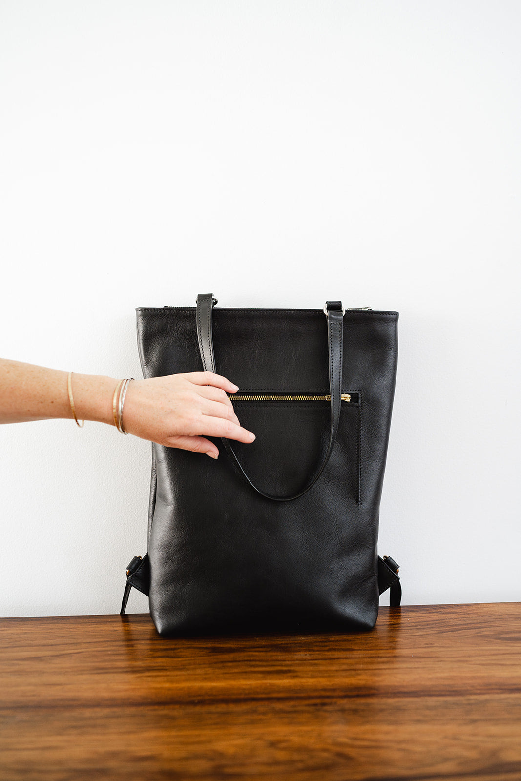 Hand reaching for The Backpack by Ella Jackson Australia – minimalist black leather backpack on timber table, showing gold zipped pocket detail.