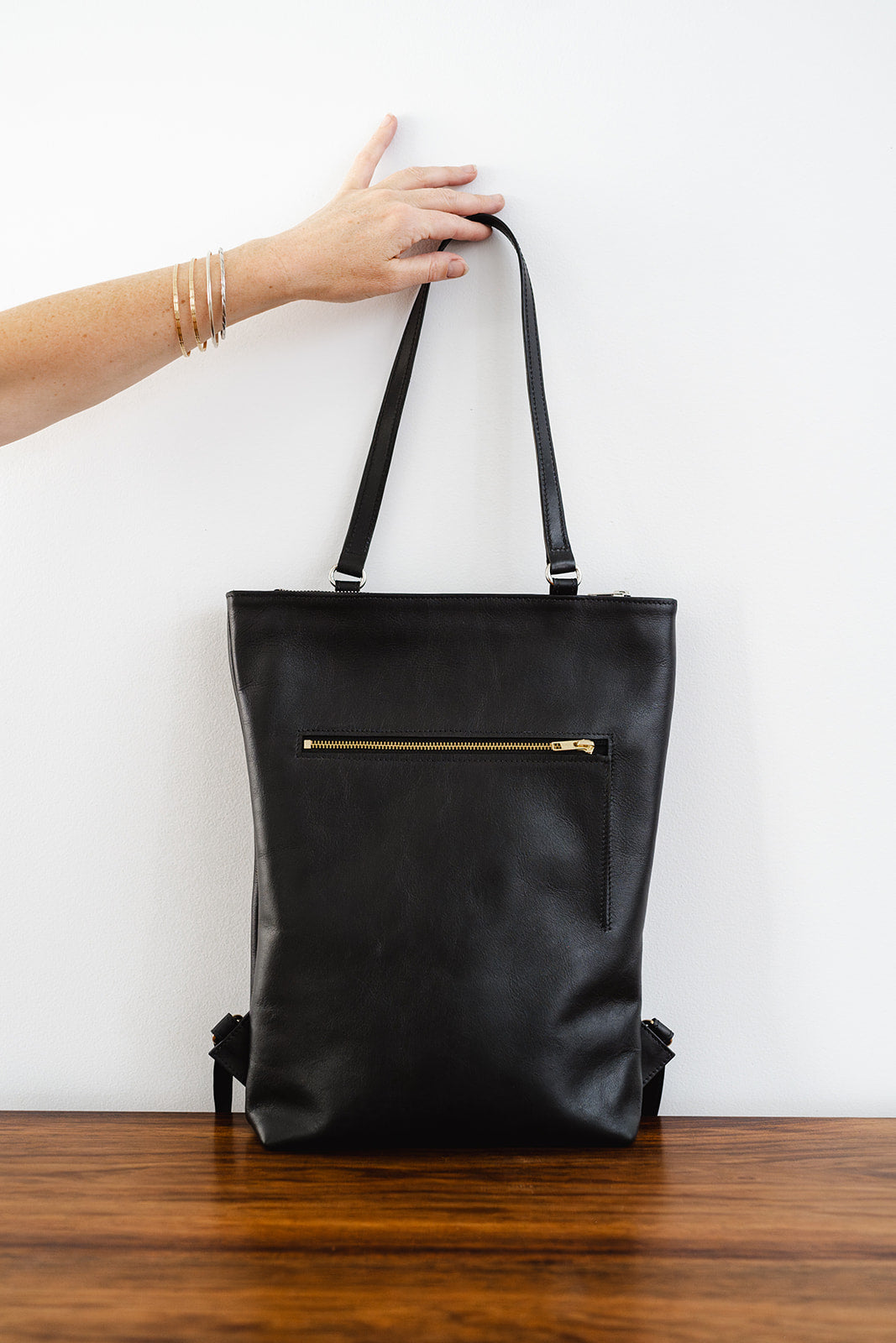 Hand lifting The Backpack by Ella Jackson Australia – black leather backpack with gold zipped pocket, minimalist styling.