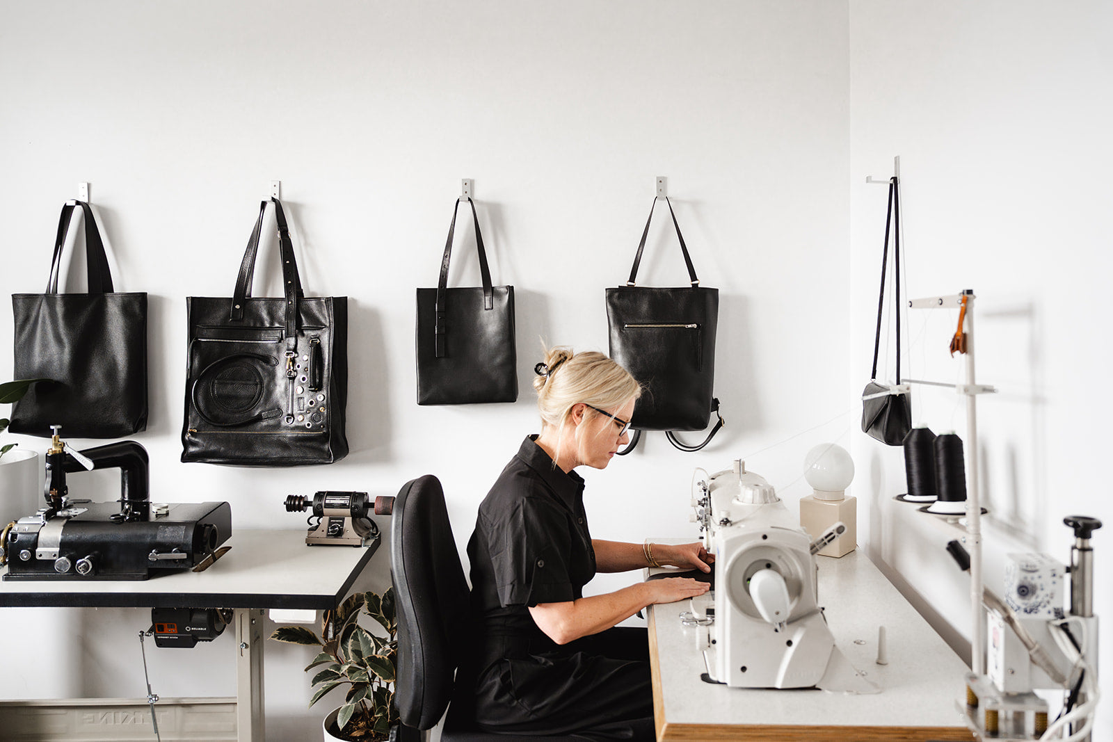 Blonde woman wearing glasses and a black outfit seated at a sewing machine in a bright, minimalist studio with black leather bags hanging on a white wall and various leatherworking tools on nearby workstations