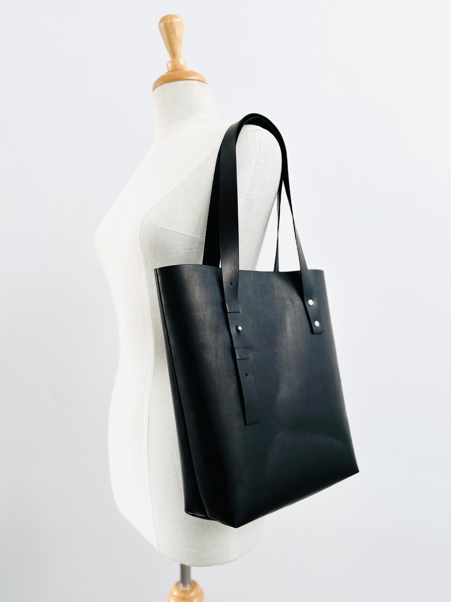 Sleek black leather tote bag elegantly draped over the shoulder of a mannequin, showcasing the bag's minimalist design and structured silhouette against a neutral background