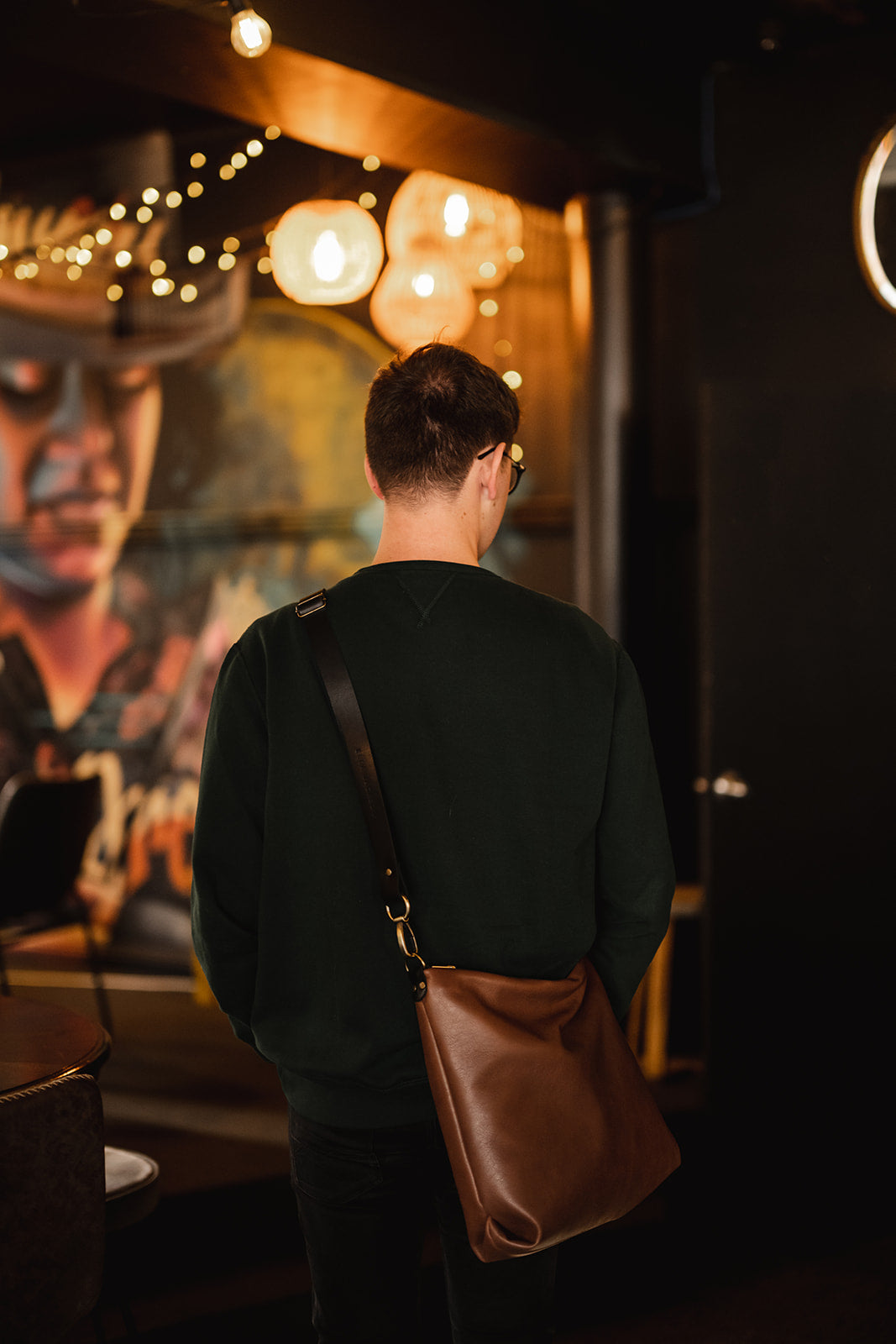 Man standing in low lit room with mural of a face on the wall. His back is turned and he is wearing a tan leather crossbody bag with black strap. The bag is the Ella Jackson Tan Leather Carryall