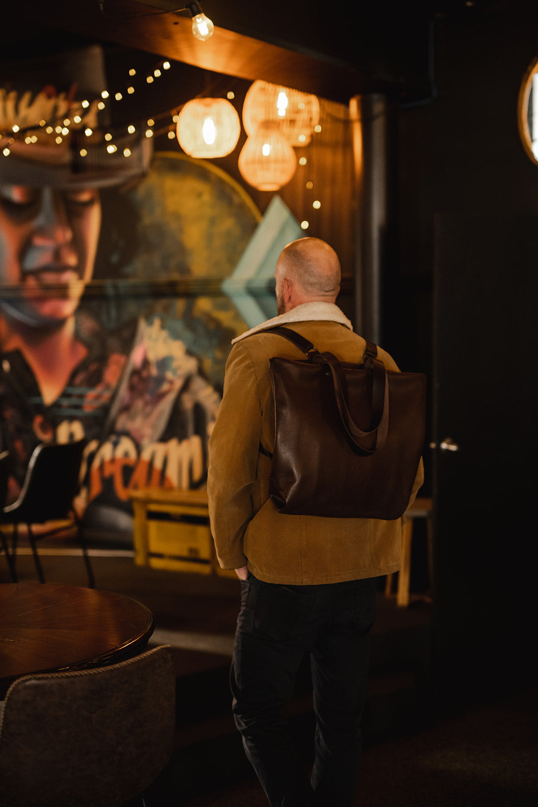 Man standing in low lit room with mural of a face on the wall. His back is turned and he is wearing a chocolate brown leather backpack. The bag is the Ella Jackson Chocolate Brown Leather Backpack & Tote