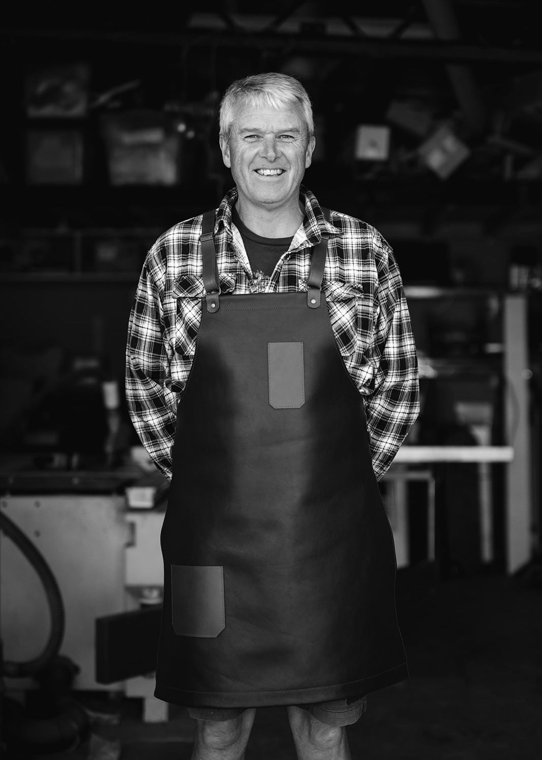 Smiling man in a workshop wearing a handmade leather apron, with a rustic background featuring various tools and equipment