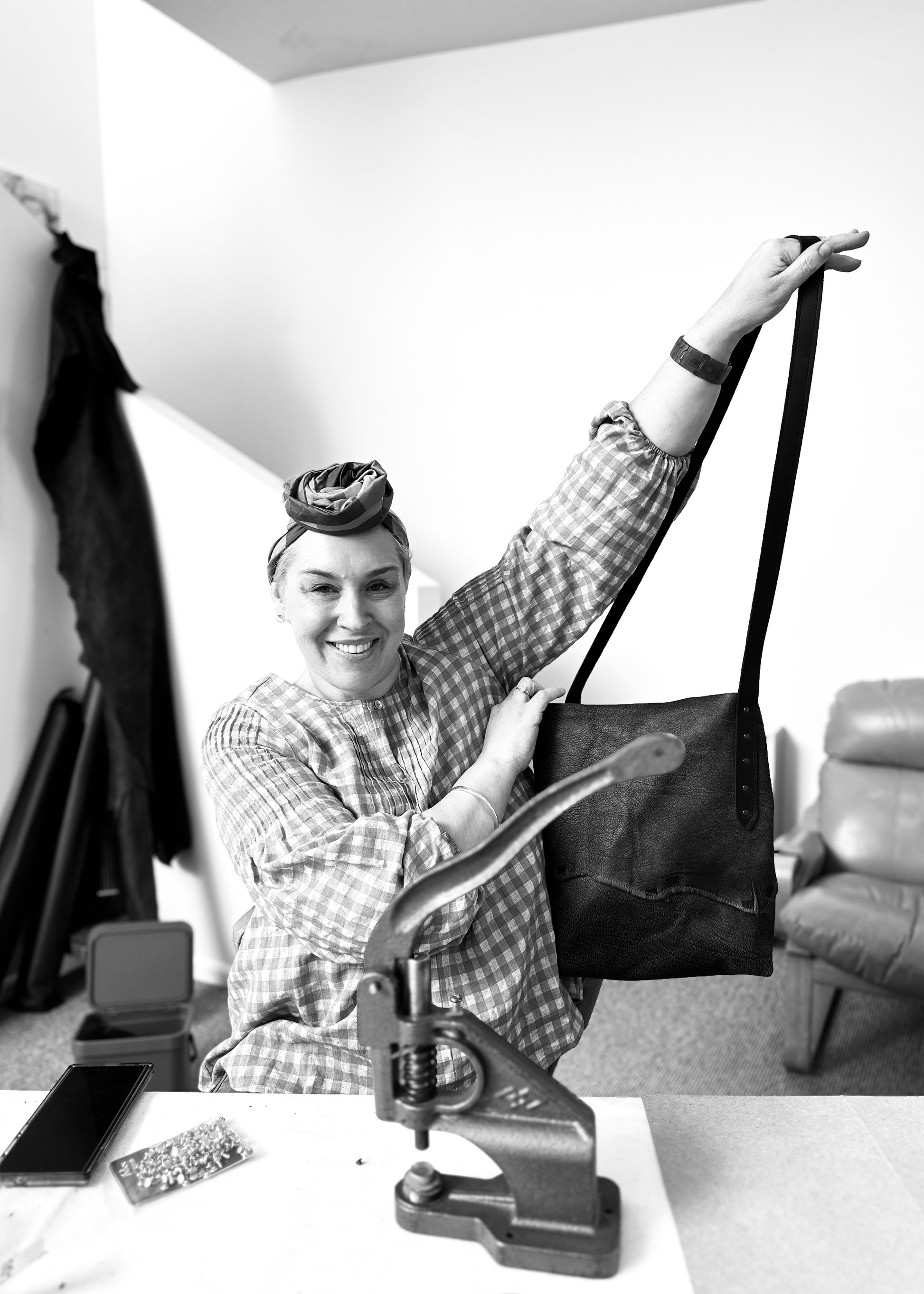 Smiling woman holding up her handmade leather tote bag during a workshop, sitting at a table with leather crafting tools, including a rivet press and metal hardware, in a creative workspace