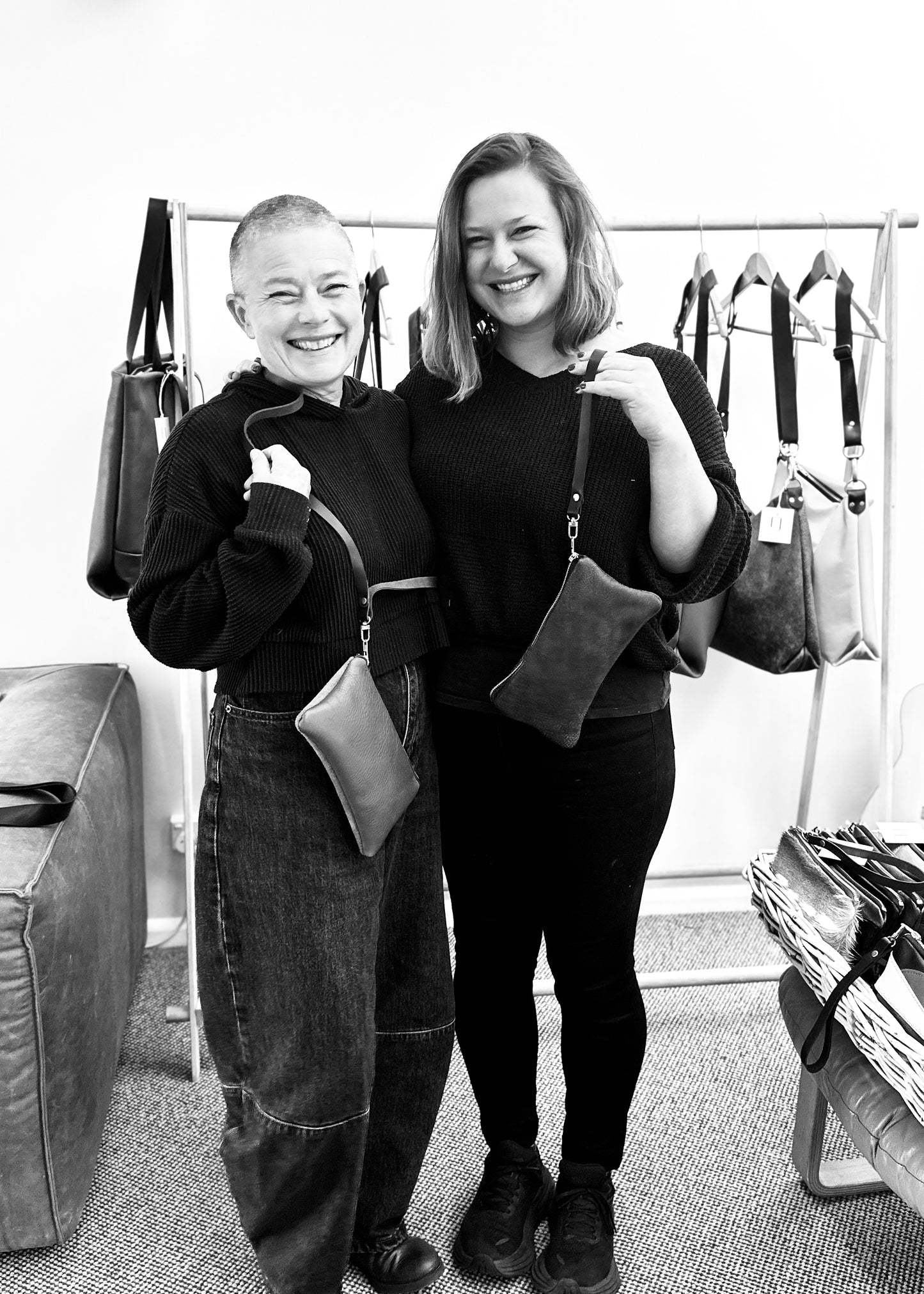 Two women smiling and holding their finished leather clutches with shoulder straps at a workshop, standing in front of a display of leather handbags. Both are dressed in cosy jumpers and exude a sense of accomplishment and joy