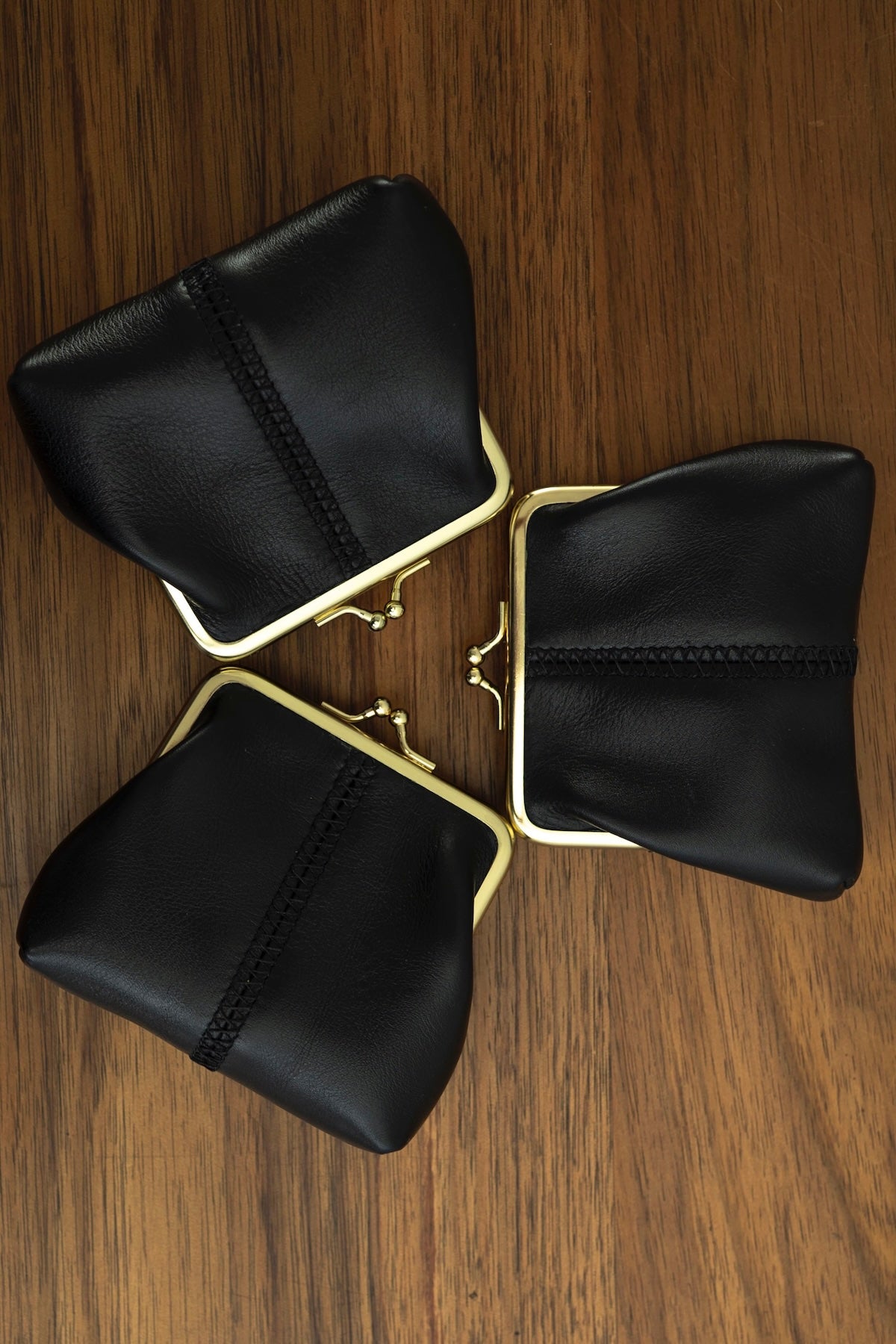 The Frame Purse by Ella Jackson Australia — three black leather frame purses arranged in a flatlay triangle on timber with gold clasps and signature central stitching
