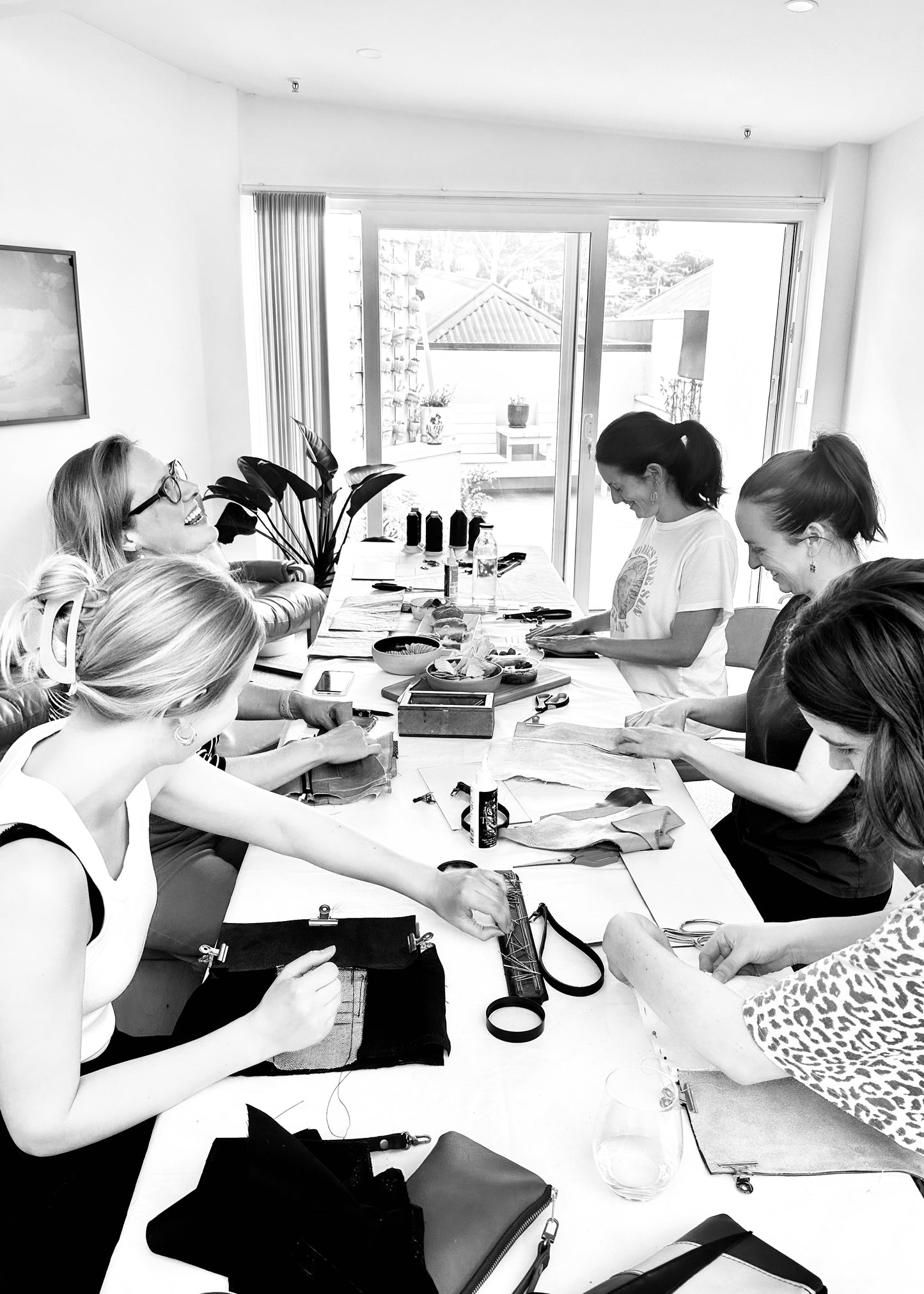 Group of women joyfully crafting leather pieces at a workshop, seated around a table covered with leather materials, tools, and finished accessories, in a bright room with natural light and greenery in the background.