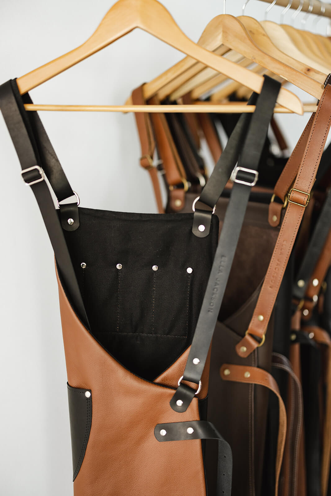 Timber rack of Ella Jackson leather aprons showing them from behind. Front apron is the Ella Jackson Jeweller Leather Apron and has a tan body, black straps and pockets, and a black cotton lining