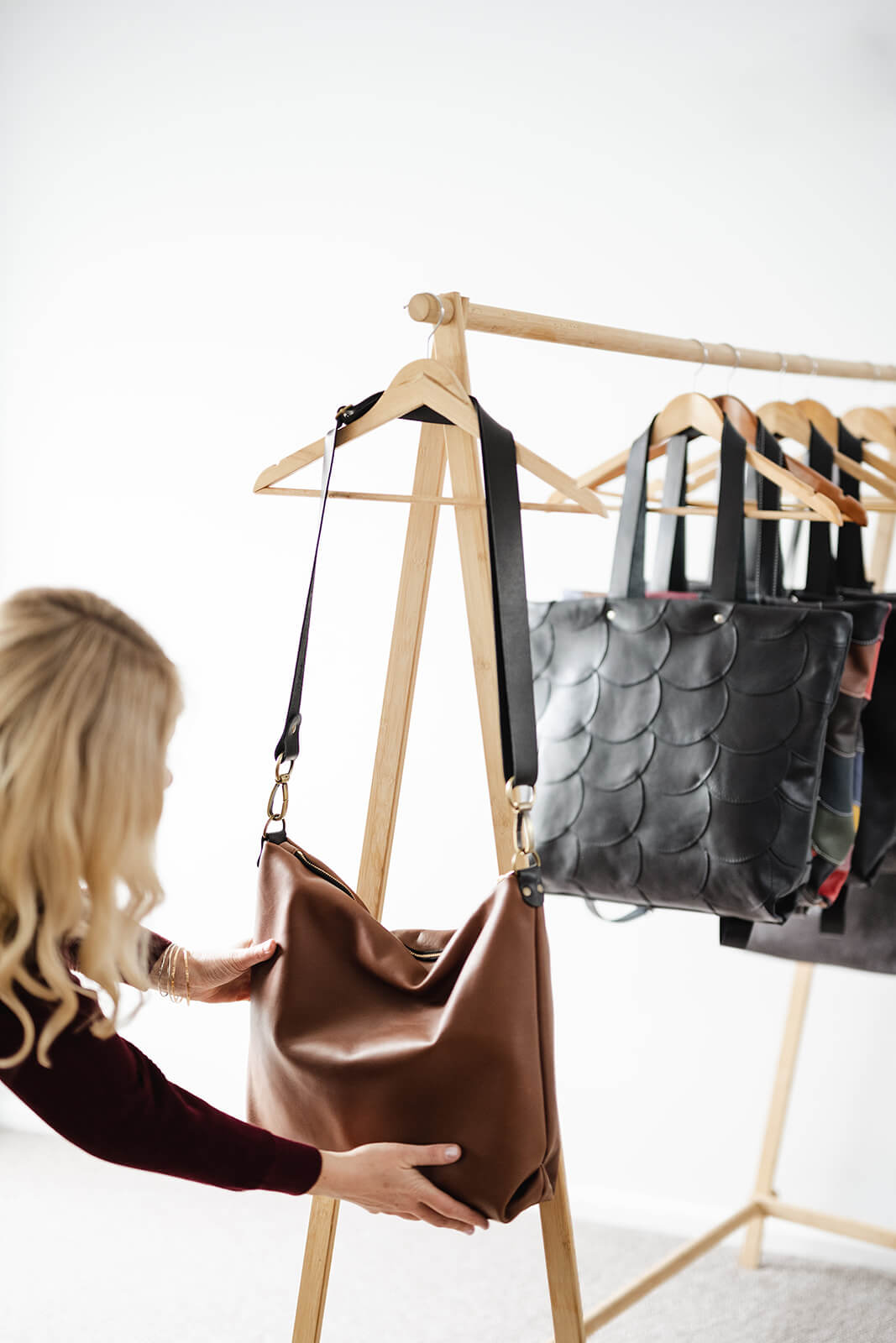Blonde woman adjusting tan leather bag hanging on a timber rack of leather bags. The bag being adjusted is the Ella Jackson Tan Leather Carryall.