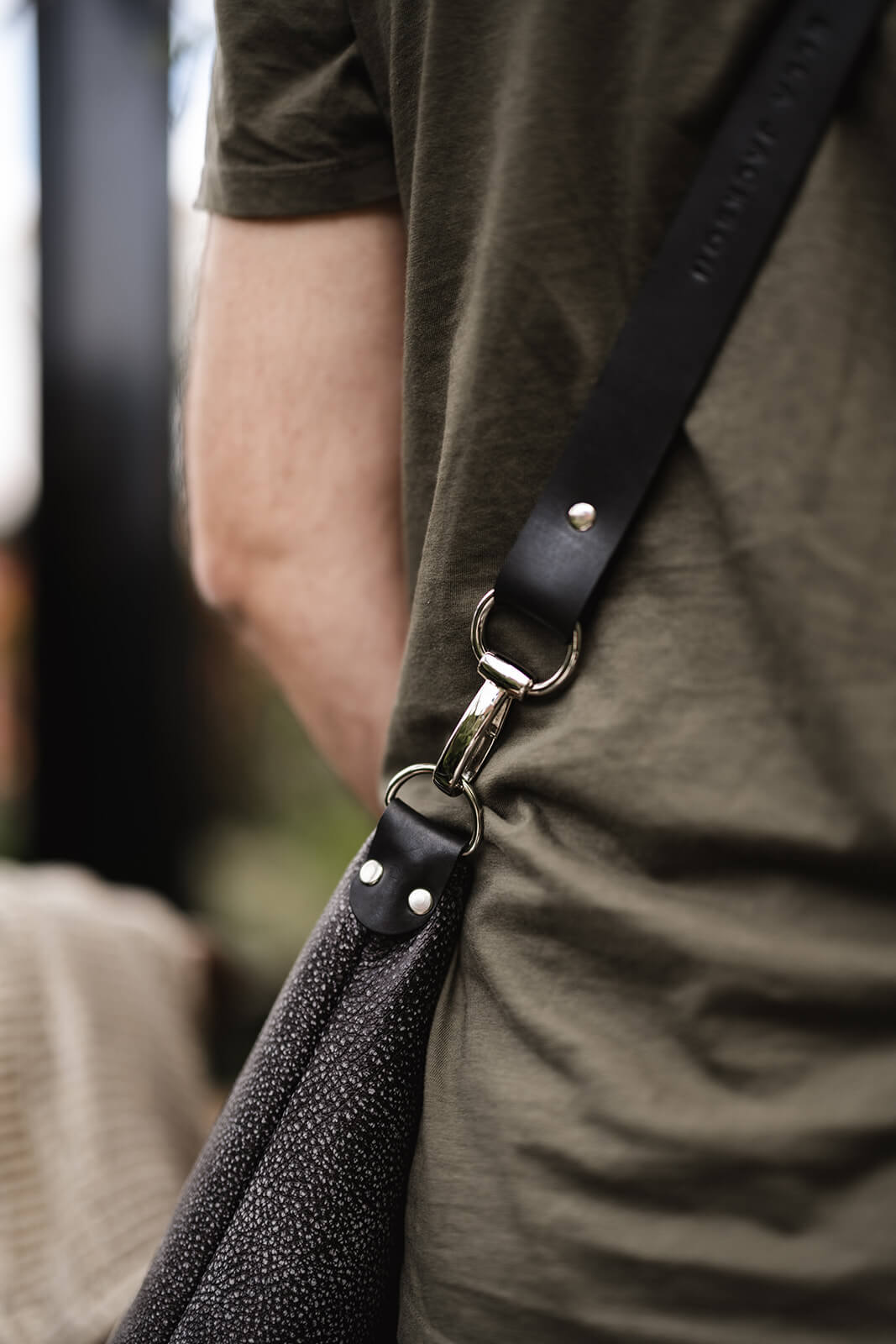 Back and arm of man wearing khaki green tshirt and a grey leather shoulder bag with black strap and silver hardware. The photo is highlighting the features of the bag. The bag is the Ella Jackson Two-tone Grey Leather Carryall