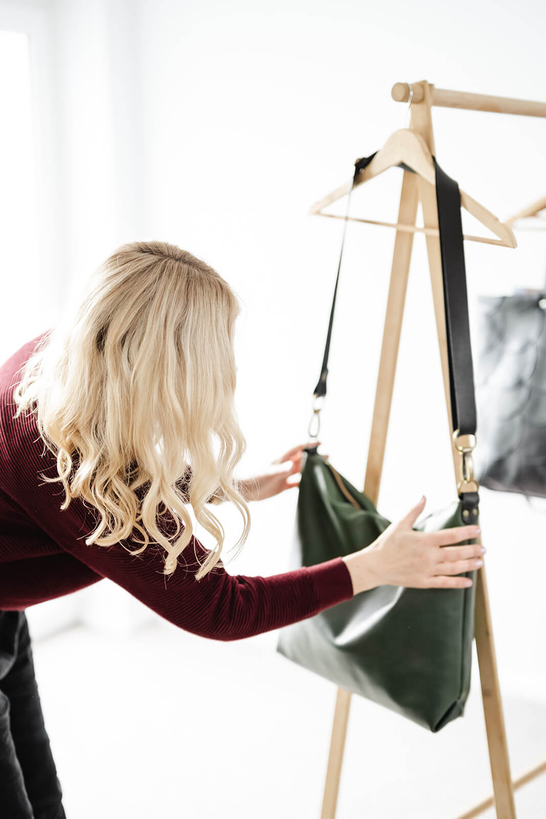 Woman arranging green leather bag on timber coat hanger. She has wavy blonde hair and a maroon jumper. The bag is the Ella Jackson Leather Carryall in Moss Green