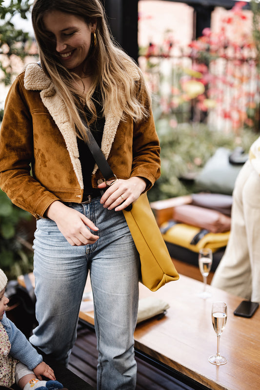 Woman smiling in sheepskin lined jacket and blue jeans and modelling a yellow leather carryall bag across body. The Ella Jackson Yellow Leather Carryall