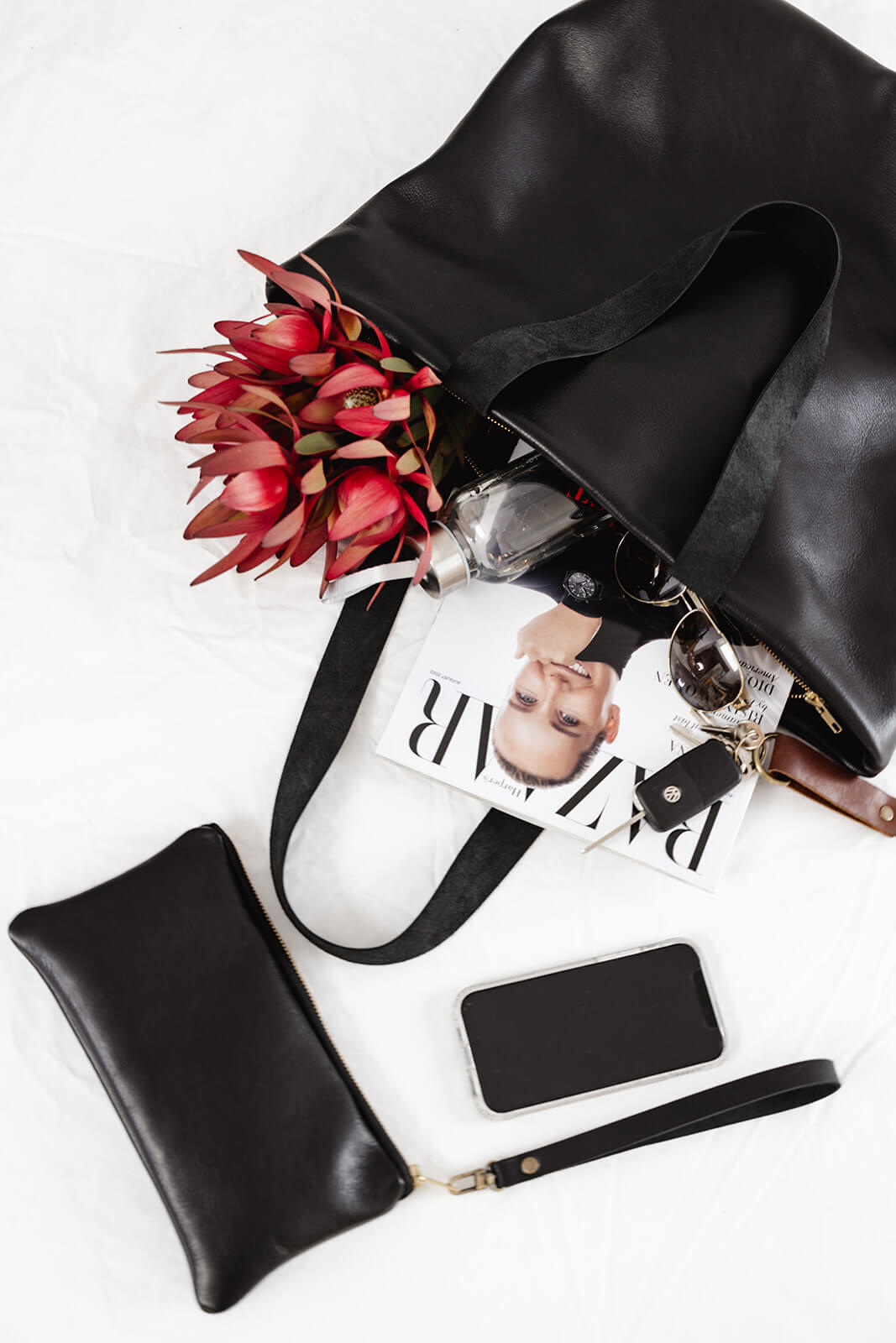 Ella Jackson Leather Backpack & Tote in Black lying on a white sheet and showing what fits in it...magazine, flowers, water bottle, glasses, keys, phone and Ella Jackson Leather Card Clutch in black