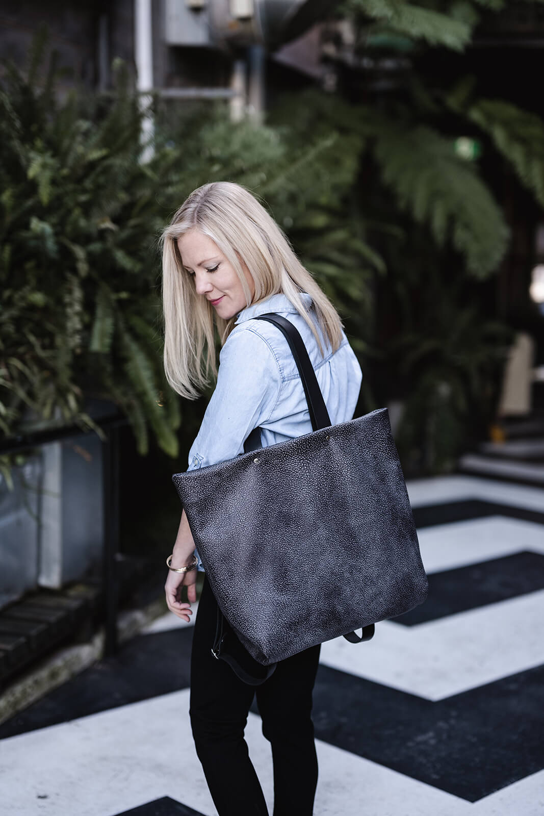 Woman with long blonde hair wearing a light denim shirt and black jeans, and looking down at her grey tote with black straps. She is standing on black and white zigzag floor in front of greenery