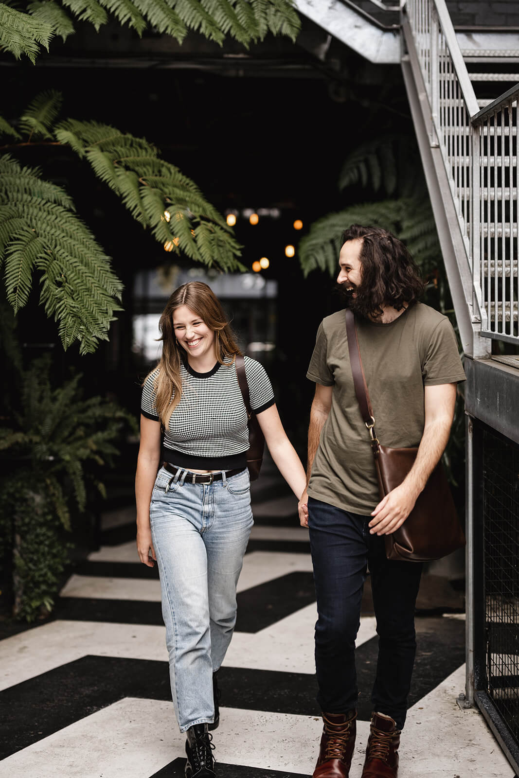 Heterosexual couple hand in hand walking towards camera and laughing. Black and white zig zag floor, ferns above. Woman is wearing black and white top and light jeans. Man is wearing green tshirt, dark jeans and brown leather boots and crossbody brown leather bag