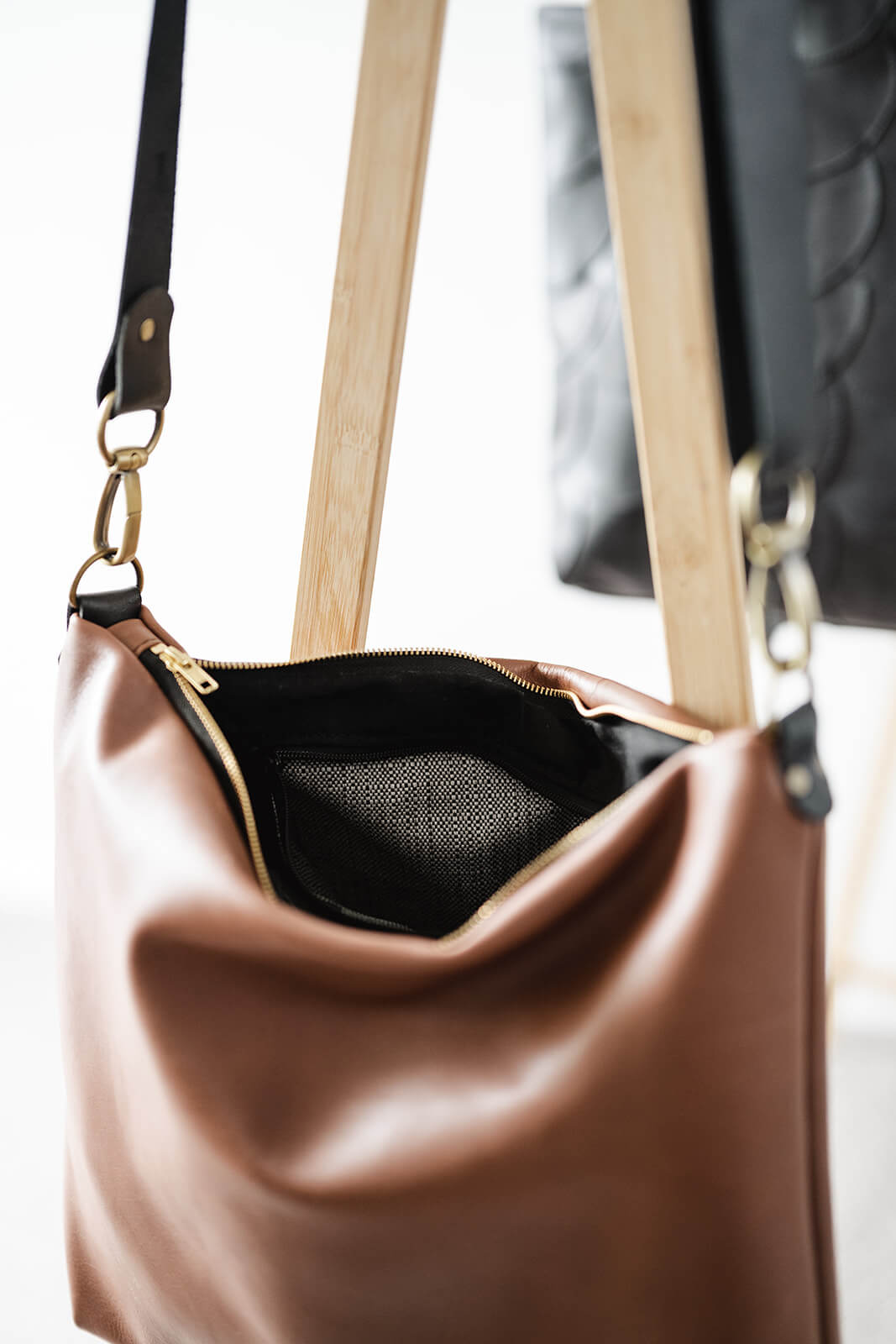 Close up product pic of a tan leather bag showing black strap, antique hardware, black and gold zip, black lining and black and white internal pocket fabric