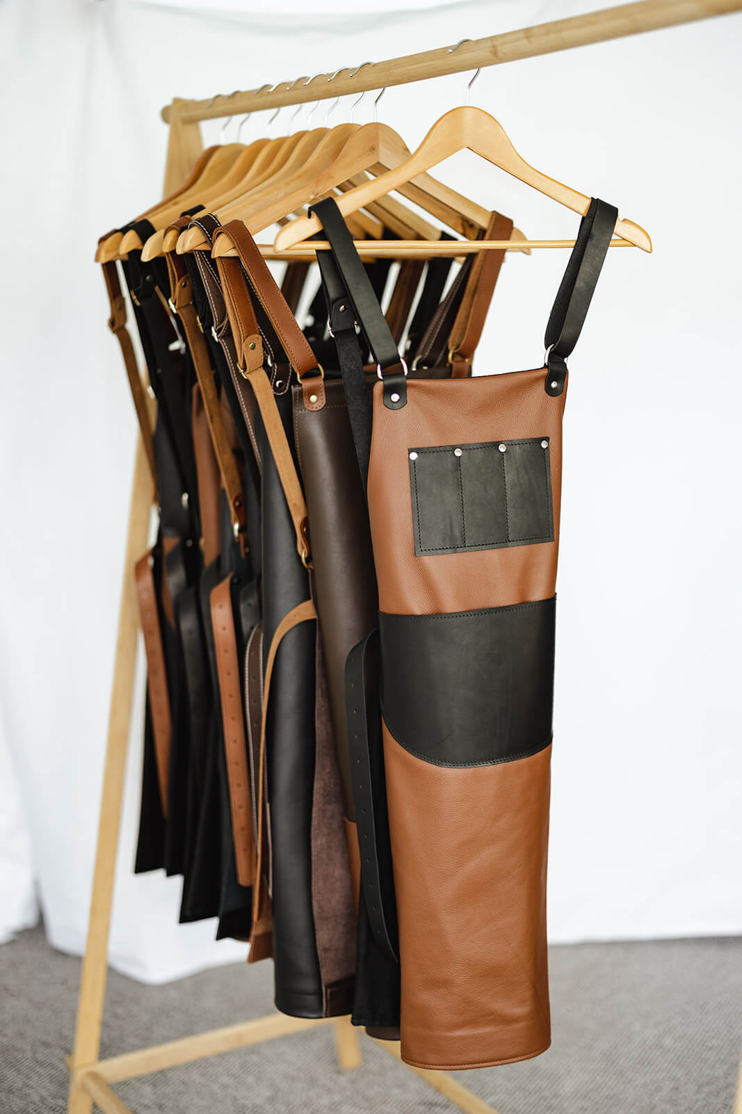 Timber rack of Ella Jackson leather aprons. Front apron is the Ella Jackson Jeweller Leather Apron and has a tan body and black straps and pockets