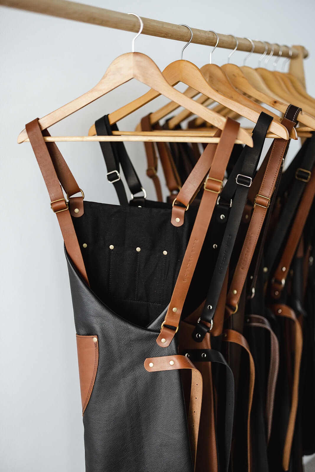 Timber rack of Ella Jackson leather aprons showing them from behind. Front apron is the Ella Jackson Jeweller Leather Apron and has a black body, tan straps and pockets, and a black cotton lining