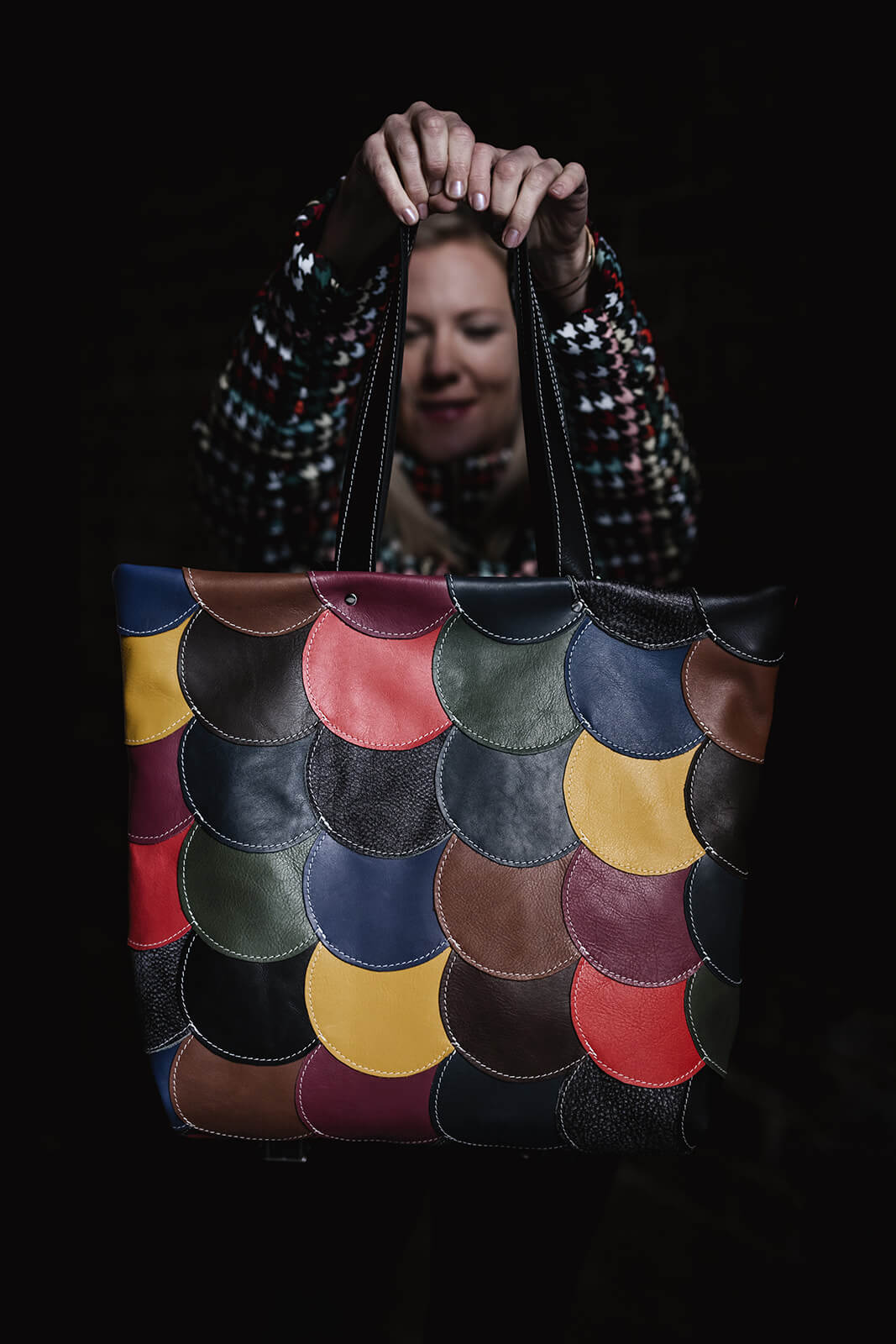 Woman in colourful jacket holding a colourful leather tote up to the camera. The tote bag is made of round pieces of leather in a lot of colours stitched together