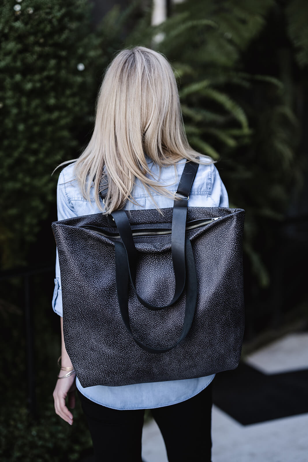 Woman standing in front of greenery and with back to camera. Blonde long hair, light denim shirt and black pants. She is modelling the Ella Jackson Two-tone Grey Leather Backpack & Tote. The bag is being worn as a backpack with black straps, silver hardware and black and silver zip. 