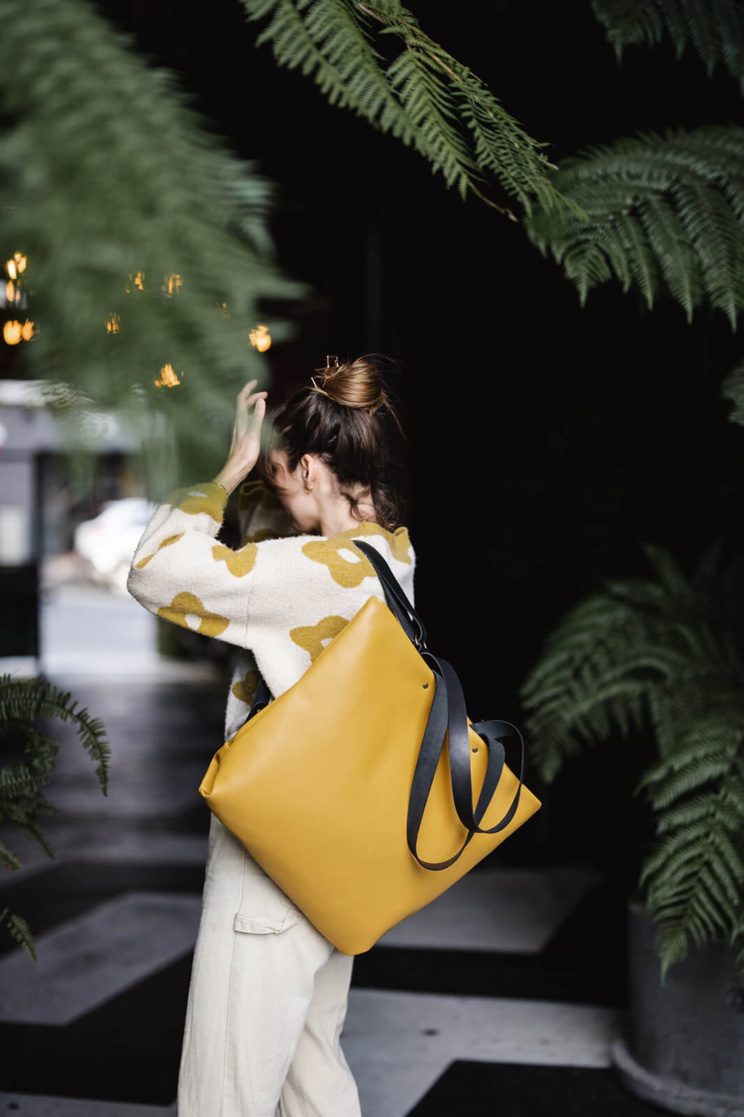 Woman standing amongst green ferns with dark background. Her hand is to her head in a modelling pose, Her clothes are beige and yellow and she is modelling a yellow leather backpack and tote as a backpack on her left shoulder. The straps are black leather