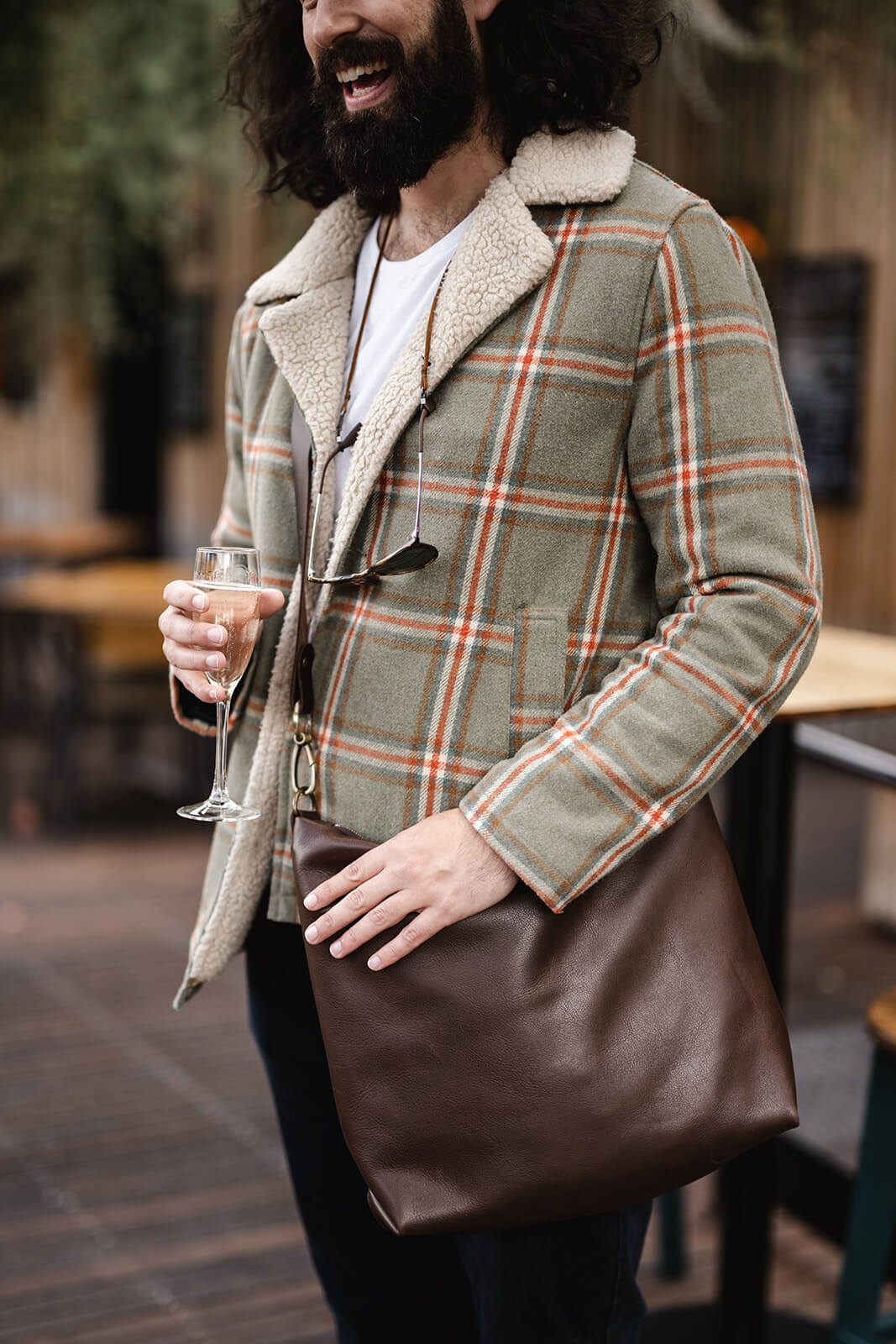 Man in Winter clothing holding a glass of champagne and a crossbody brown leather bag in the other. The bag is the Ella Jackson Chocolate Brown Leather Carryall