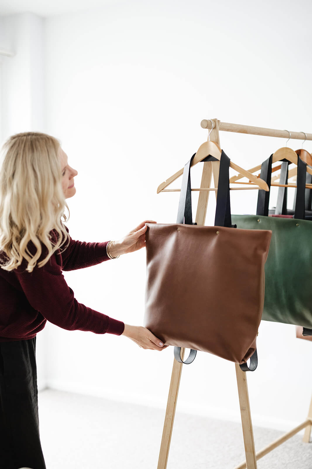 Tan Leather Backpack & Tote in Ella Jackson brand hanging on timber clothes rack as a tote and showing black leather tote straps. Designer and Maker, Ella Jackson standing behind adjusting bags on hooks and wearing black pants and a maroon jumper with wavy blonde hair