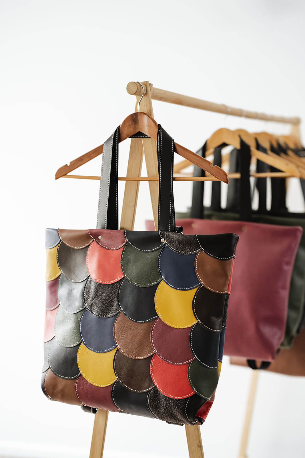 Timber rack with timber coat hangers and white background. A bag is hanging on the front. It is a tote bag made of multiple round leather circles stitched together. The circles are all different colours