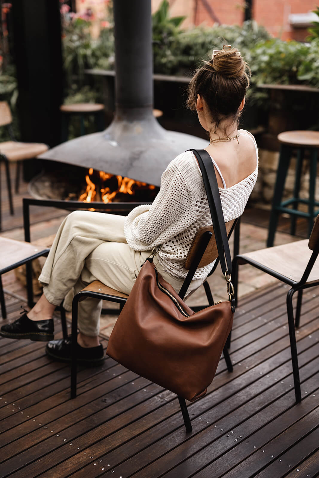 woman sitting on chair and facing open fire. Tan leather bag with black strap hanging on back of chair. The bag is the Ella Jackson Tan Leather Carryall
