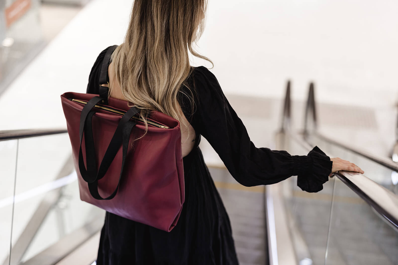 Woman in long black dress and cherry leather backpack riding down an escalator. The backpack is the Cherry Leather Backpack & Tote by Ella Jackson