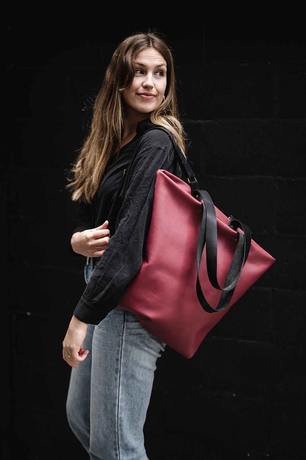 Woman in black top, pale blue jeans, and cherry leather backpack on one shoulder. She is looking over her shoulder and has long flowing hair. The backpack is the Cherry Leather Backpack & Tote by Ella Jackson