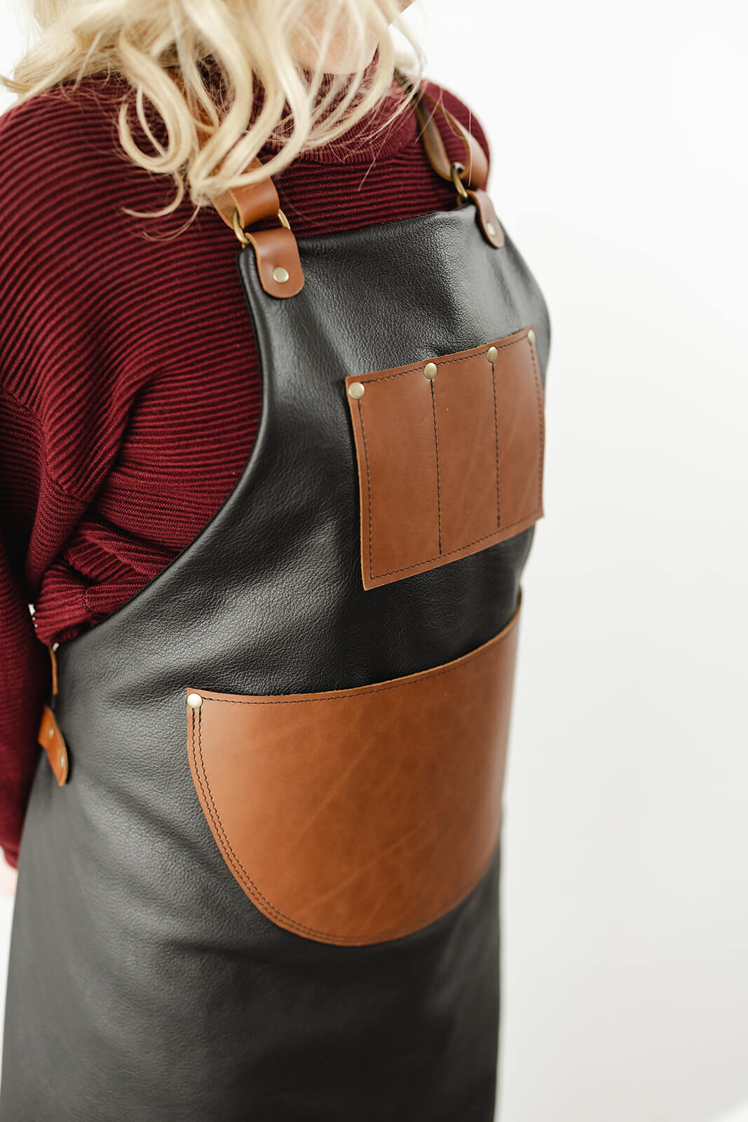 Woman with blonde hair and maroon jumper wearing a black and tan leather apron. The apron has a black leather body, tan straps, a tan leather chest pocket with 3 compartments and a large half circle tan leather waist pocket. It is the Ella Jackson Jeweller Leather Apron