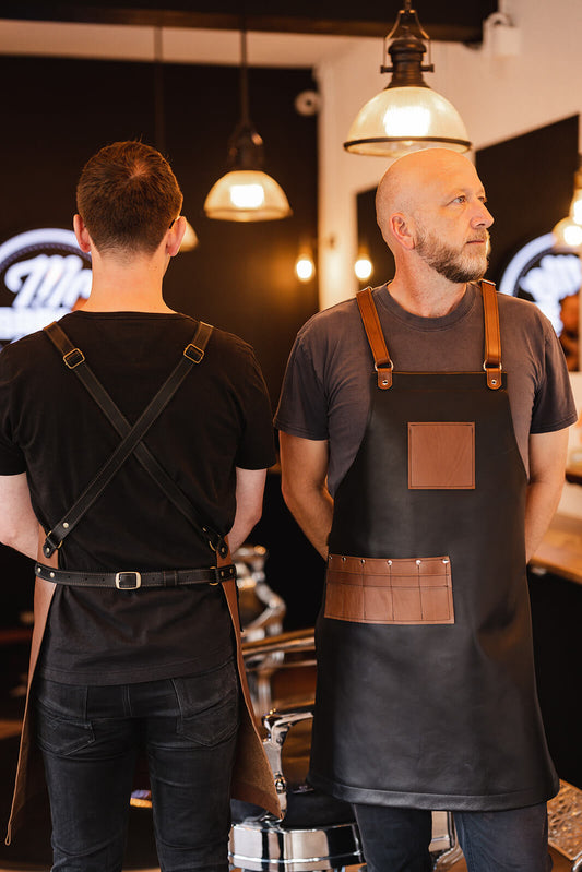 Barber on left with back turned and wearing Ella Jackson leather apron with tan body and black straps. Barber on right facing forwards wearing Ella Jackson leather apron with black body and tan leather pockets and straps