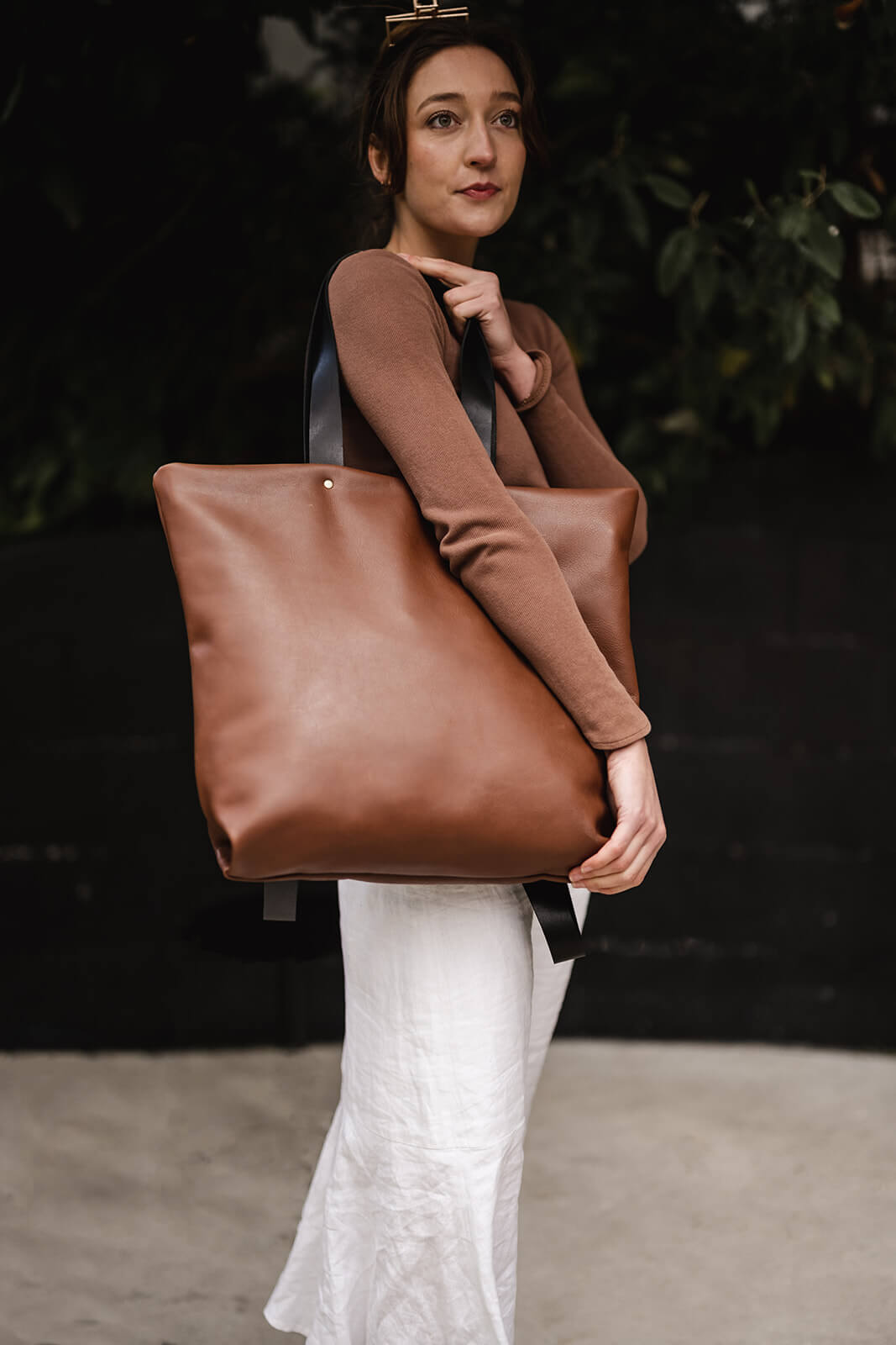 Glamorous woman in brown top and white skirt staring into the distance and wearing tan leather tote with black straps. The tote is the Tan Leather Backpack & Tote by Ella Jackson