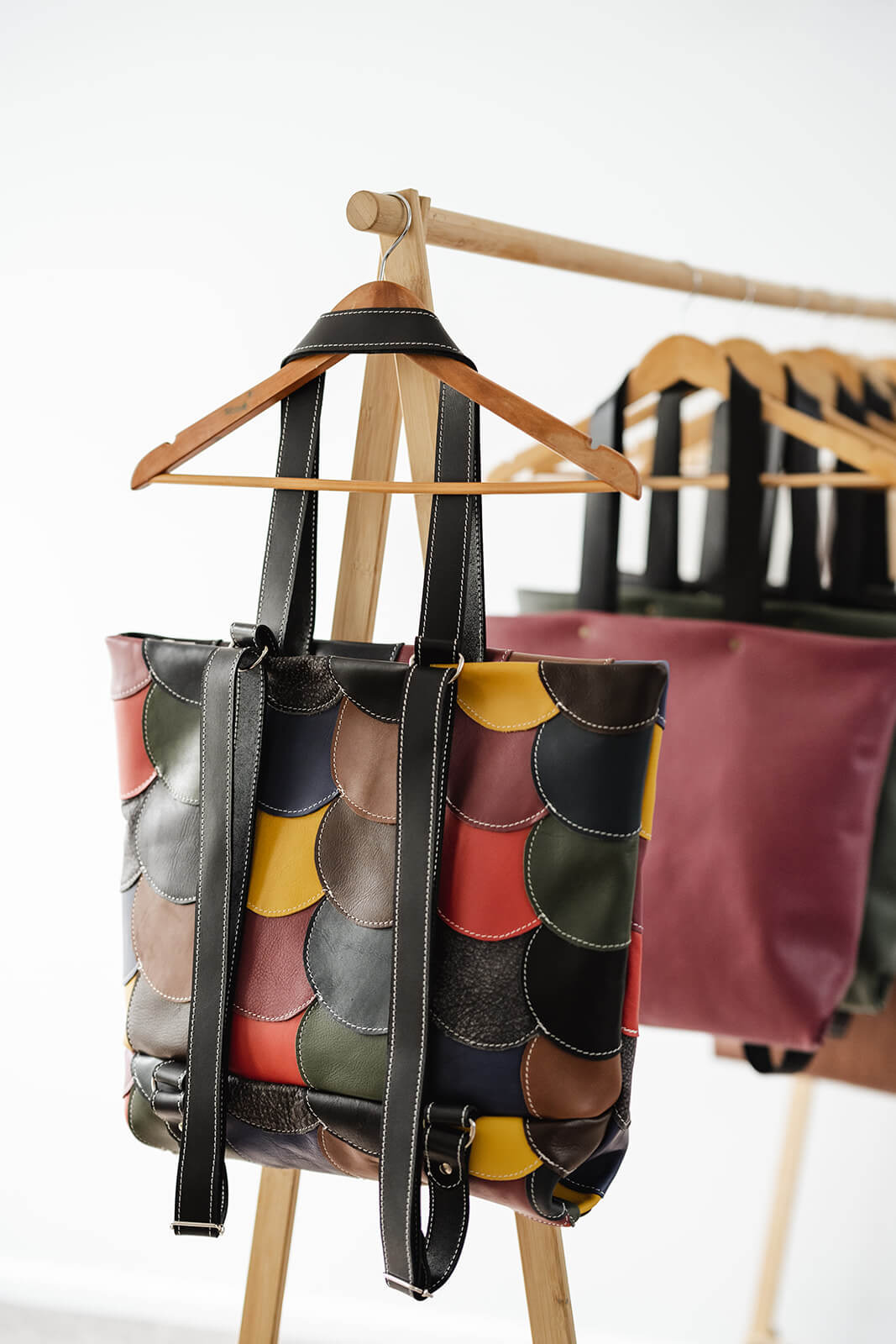 Timber clothes rack with white background. The front coat hanger has a colourful leather backpack and tote hanging as a tote and showing the backpack straps at the back. The bag is made of small circles of different coloured leather circles stitched together. It is The Circle Bag by Ella Jackson in Multicolour