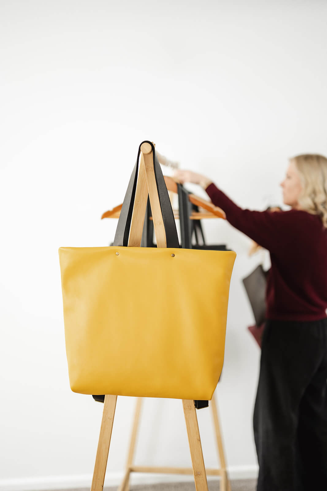 Yellow Leather Backpack & Tote in Ella Jackson brand hanging on timber clothes rack as a tote and showing black leather tote straps. Designer and Maker, Ella Jackson standing behind adjusting bags on hooks and wearing black pants and a maroon jumper with wavy blonde hair