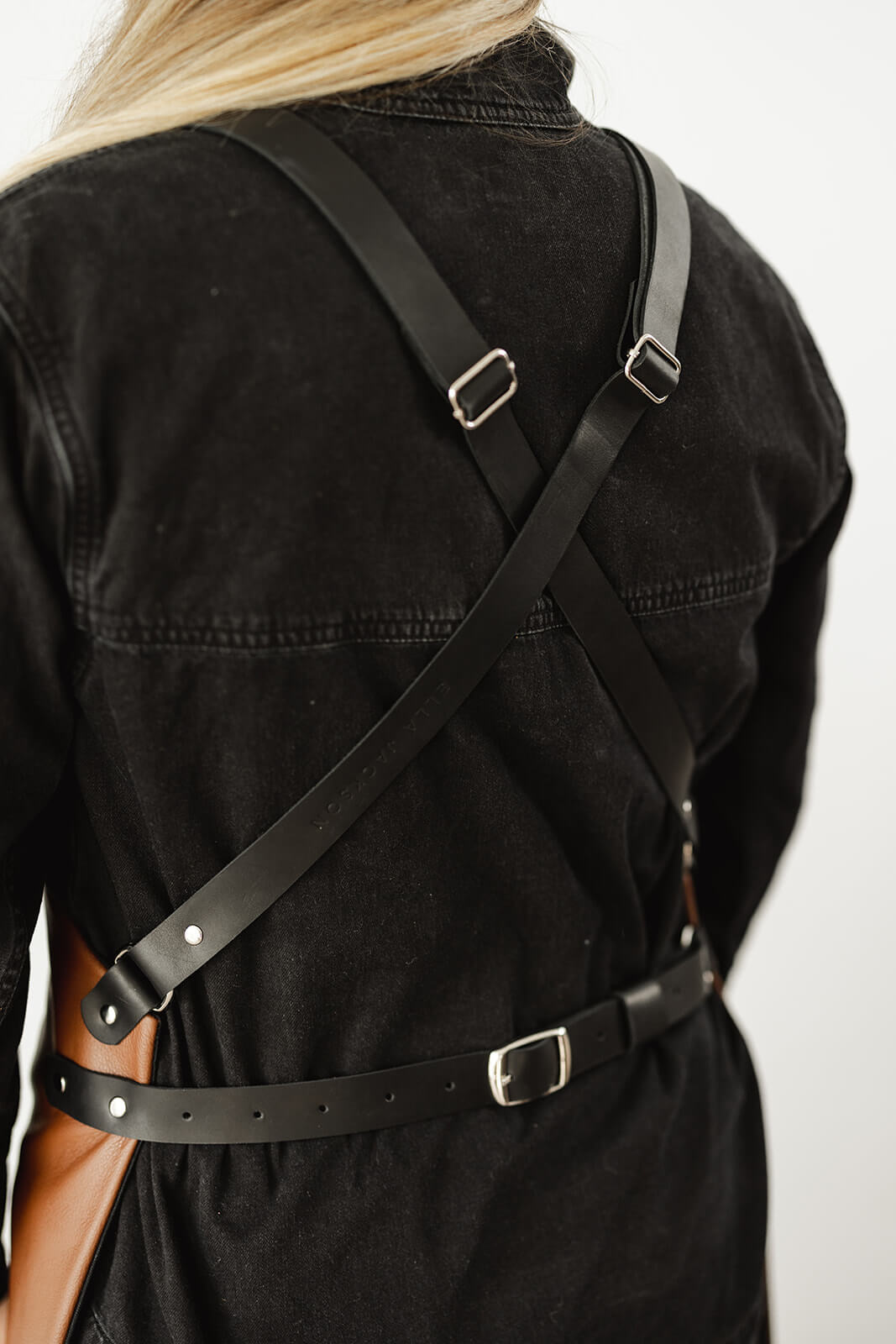 Back view of woman with black shirt wearing a leather apron. The pic shows the strap configuration from the back...black leather crisscross shoulder straps, a waist strap and silver hardware