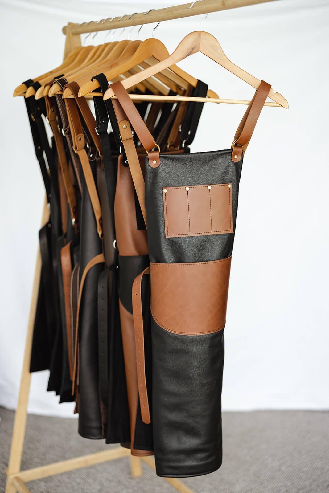 Timber rack of Ella Jackson leather aprons. Front apron is the Ella Jackson Jeweller Leather Apron and has a black body and tan straps and pockets