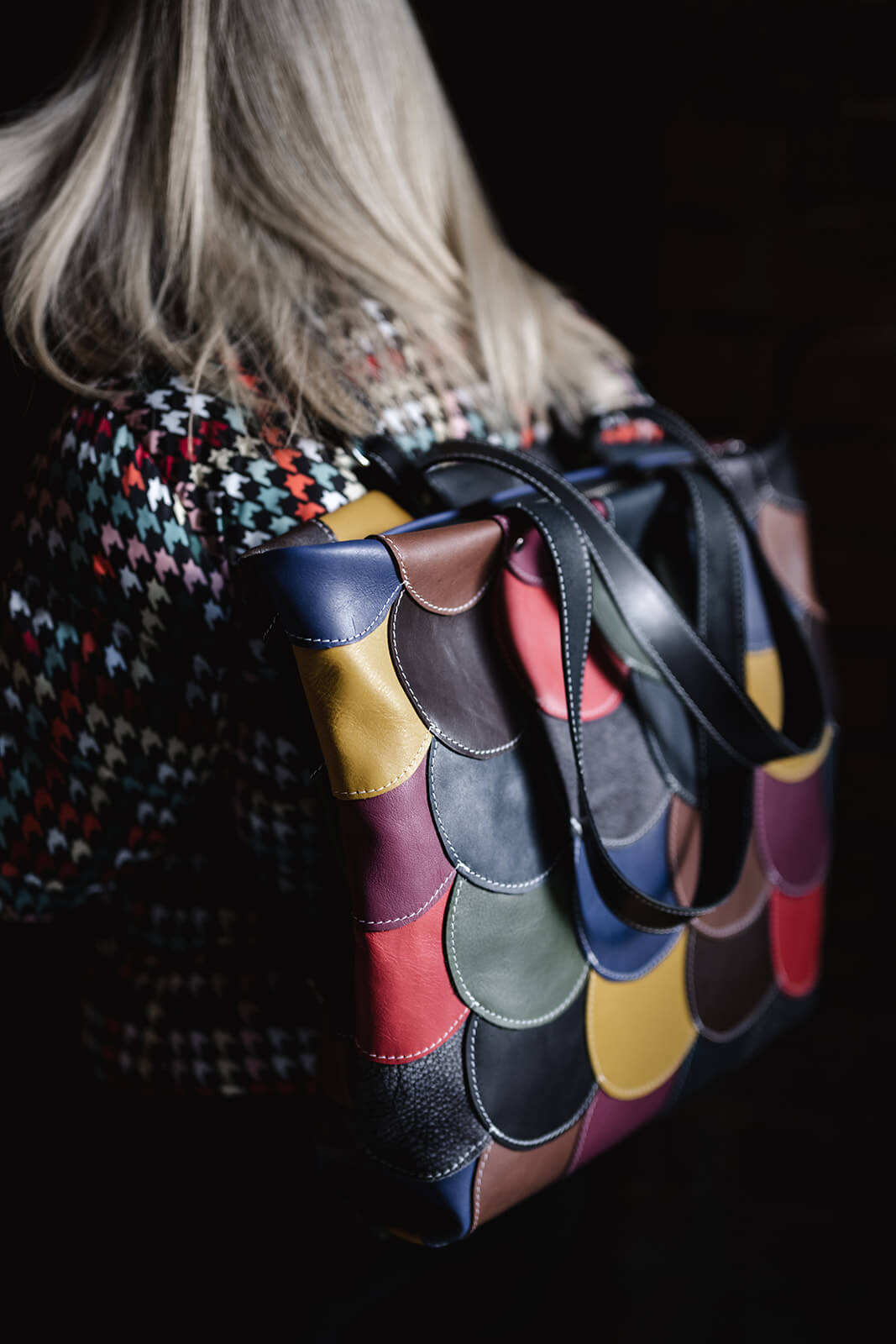 Back of a woman with straight blonde hair and a colourful puffer jacket wearing a multicoloured leather backpack. The backpack is made of leather rounds in many colours.