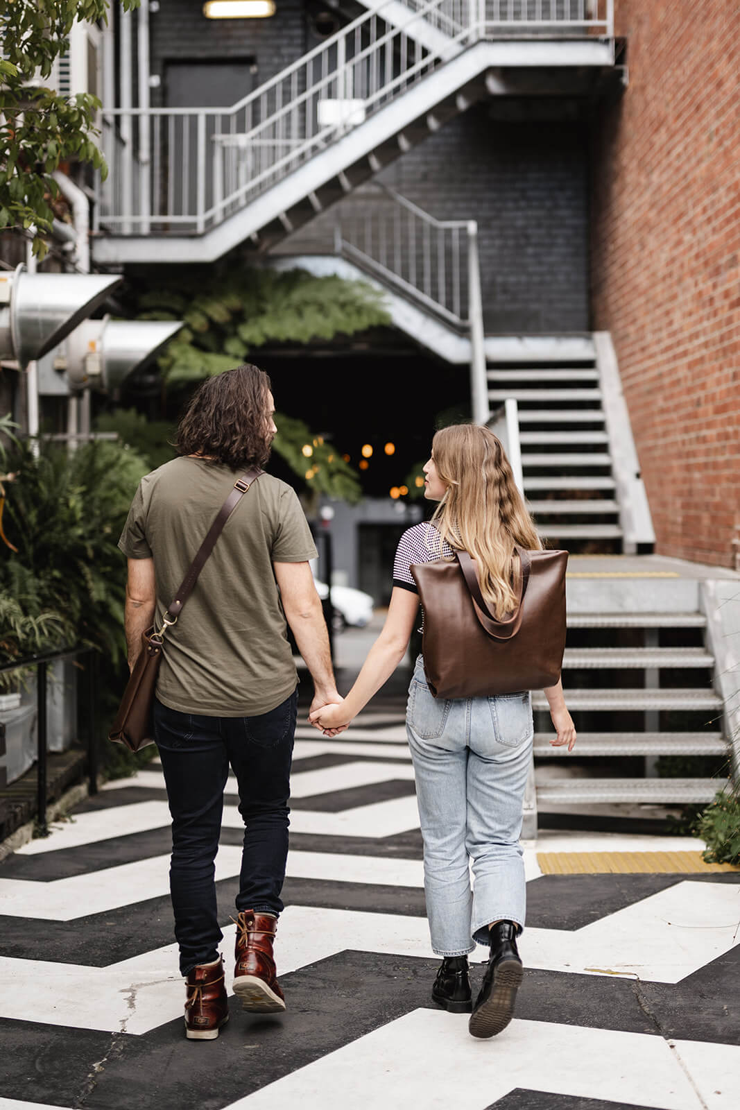 A man and woman walking hand in hand (interesting staircase behind). They are modelling Ella Jackson leather bags. The man on the left is wearing a brown leather crossbody bag and the woman on the right is wearing a brown leather backpack.