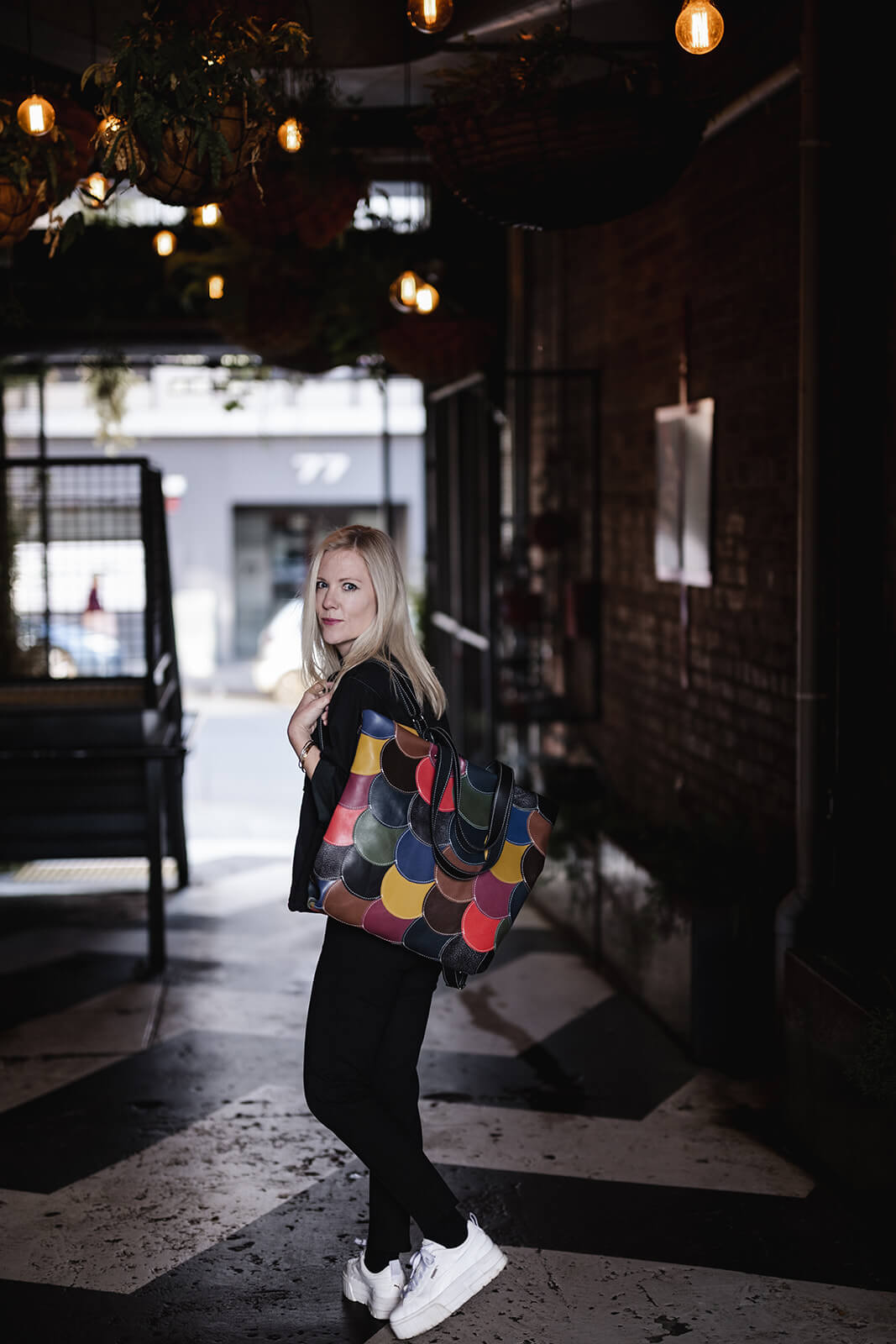Woman standing in an alley. She has straight blonde hair and is wearing black top and pants, white shoes and a multicoloured backpack. She is facing away but turning her head over her shoulder that the backpack is on. The backpack is made of colourful rounds of leather in lots colours all stitched together. It is The Circle Bag in Multicolour by Ella Jackson