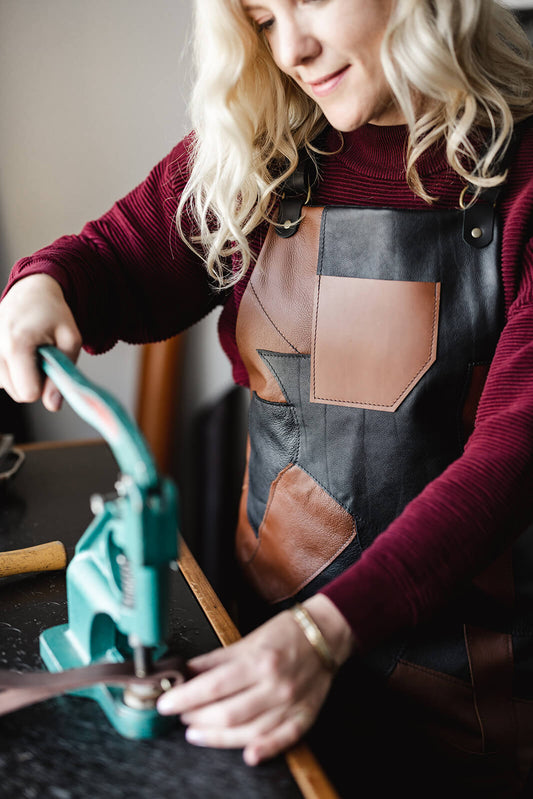 Woman with blonde wavy hair, maroon jumper and a black and tan leather apron using a rivet press at a work bench. The apron is The Maker Leather Apron by Ella Jackson