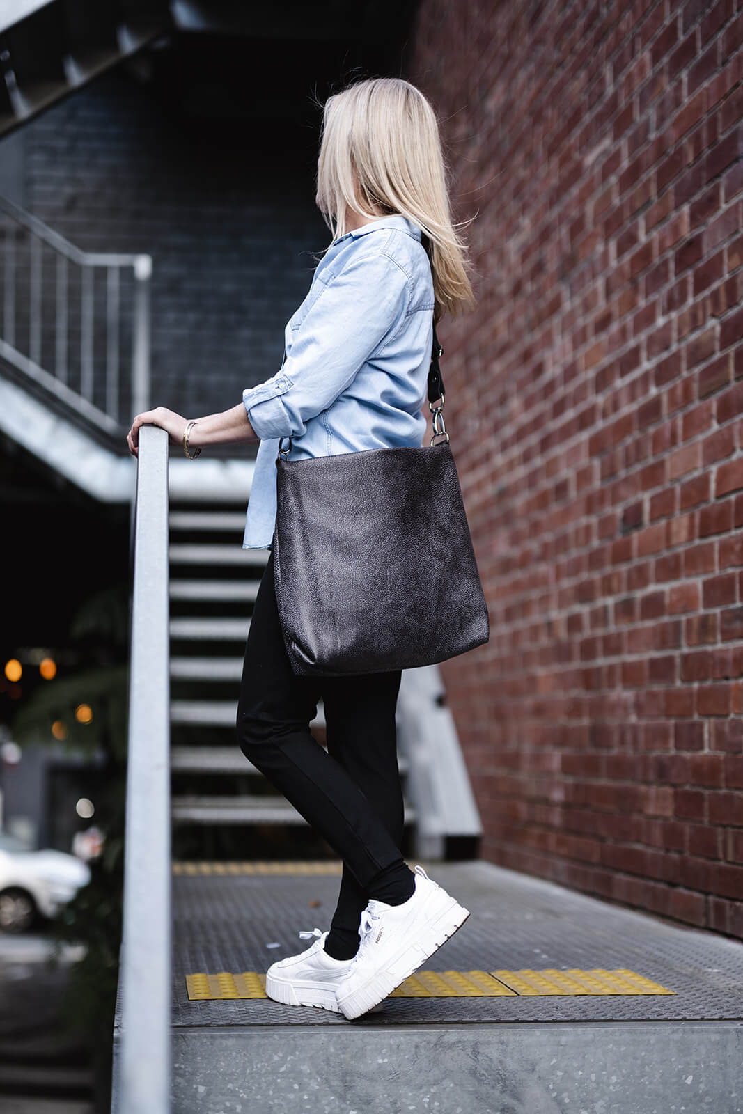 Lady standing on external stairs holding onto rails with profile view. Blonde hair, denim shirt, black jeans, white shoes and holding onto rail. Lady is modelling grey crossbody bag with black strap. The bag is the Ella Jackson Two-tone Grey Leather Carryall