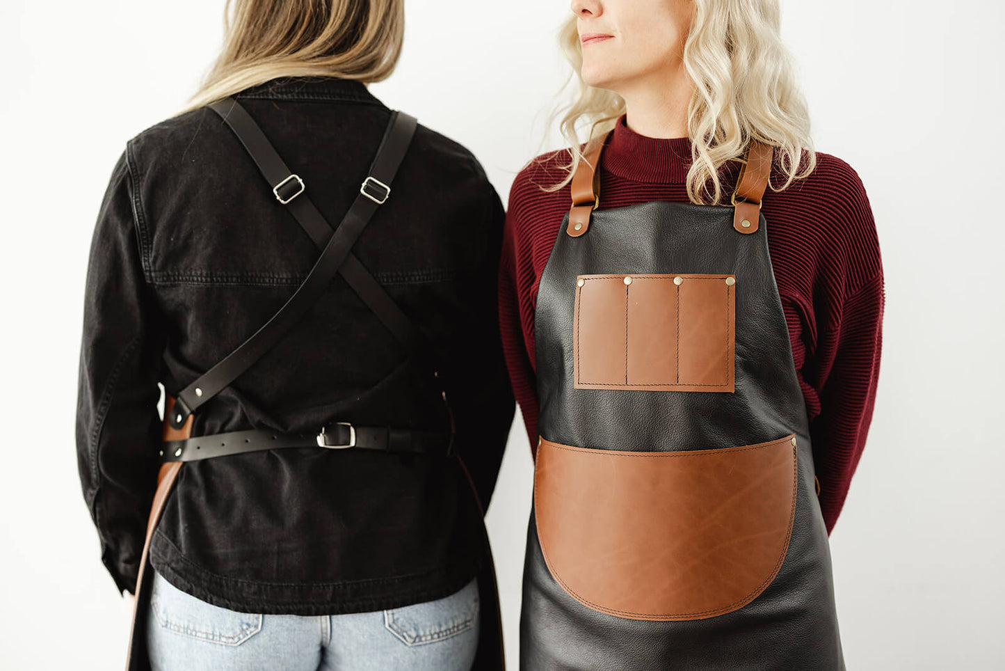 Product shot showing back and front of the Ella Jackson Jeweller Leather Apron. Woman on left is showing black leather crisscross shoulder and waist straps; woman on front is showing front of apron with black leather body, tan chest and waist pocket and tan leather straps