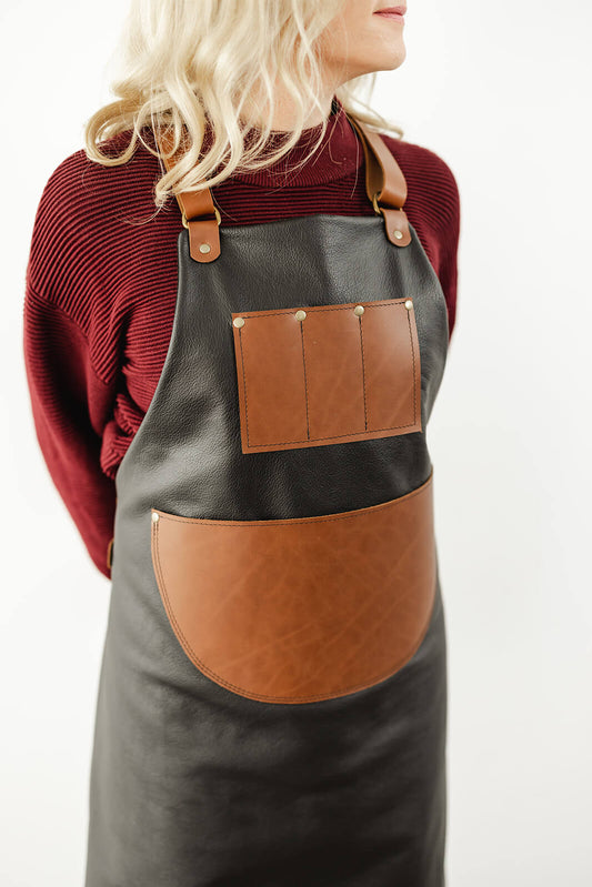 Woman with blonde hair and maroon jumper wearing a black and tan leather apron. The apron has a black leather body, tan straps, a tan leather chest pocket with 3 compartments and a large half circle tan leather waist pocket. It is the Ella Jackson Jeweller Leather Apron
