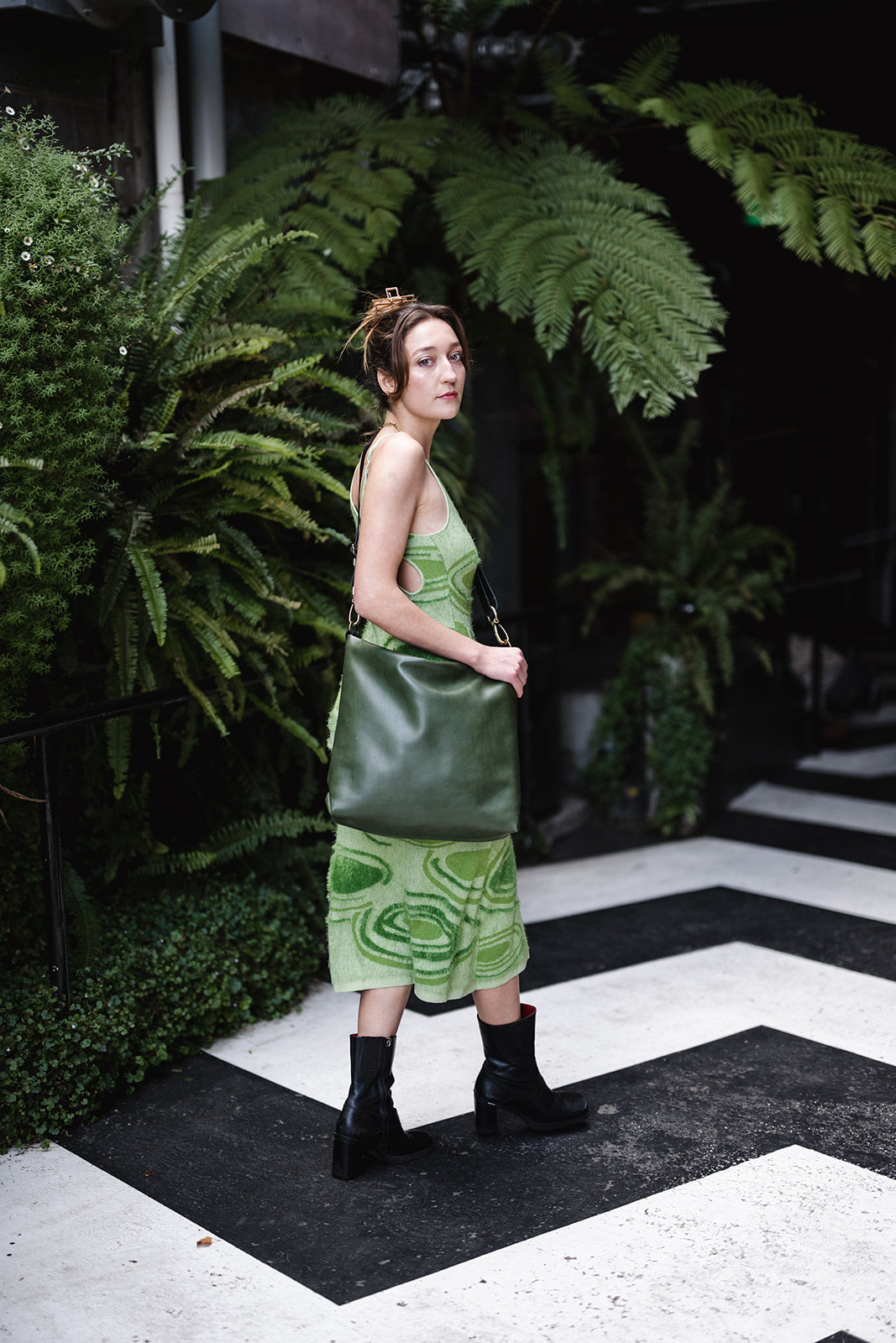 Woman in sleeveless green dress standing amongst ferns and modelling green leather bag. The bag is the Ella Jackson Leather Carryall in Moss Green
