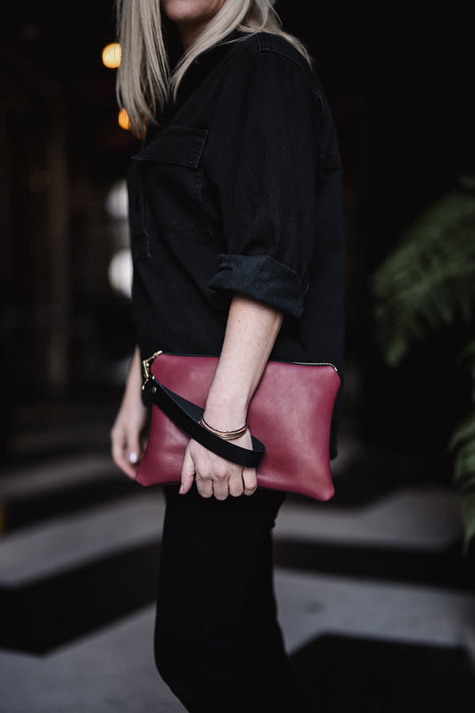 Woman dressed in black and holding a cherry leather clutch with black strap. The clutch is the Ella Jackson Oversized Leather Clutch in Cherry 