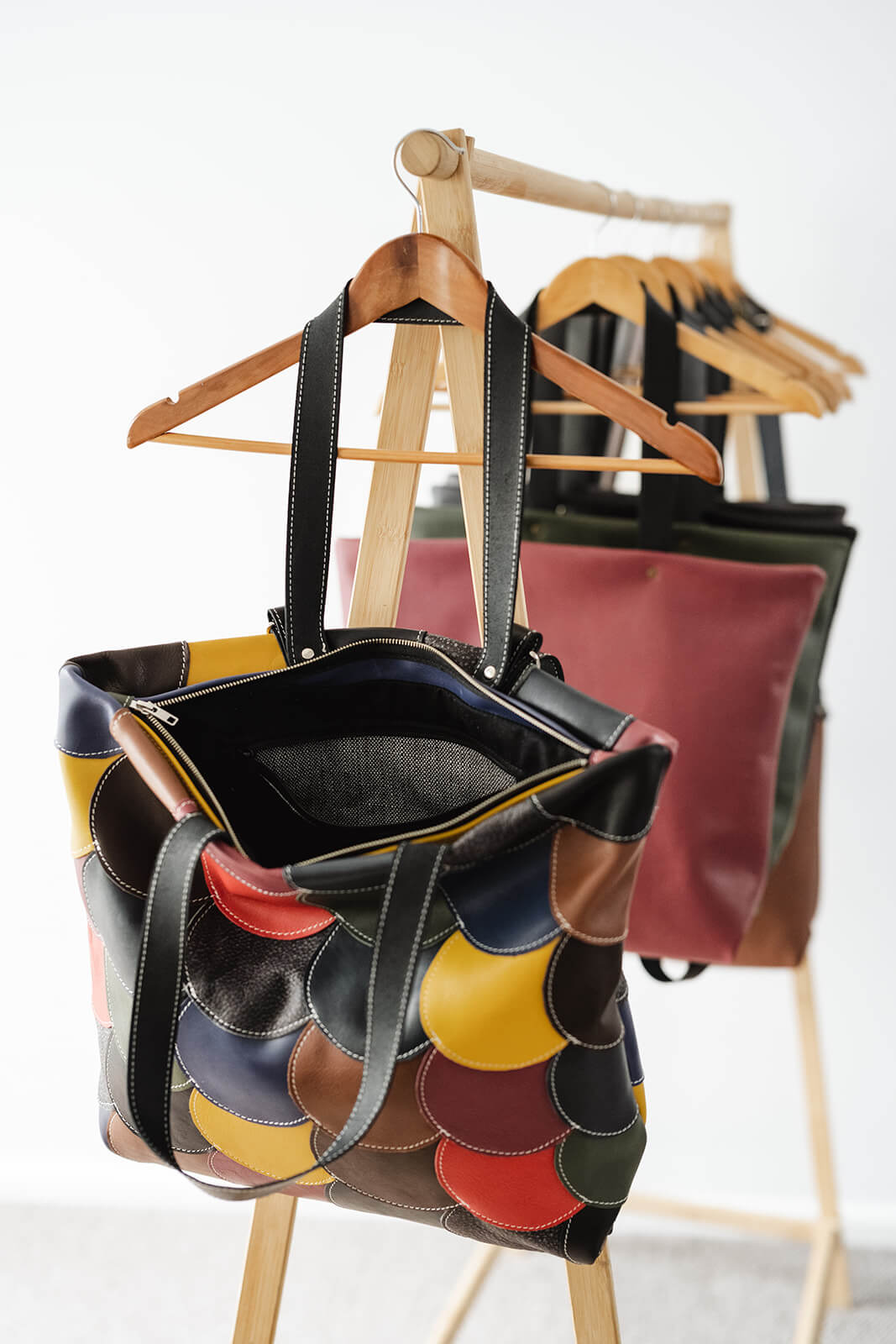 Timber rack in front of white wall with timber coat hangers holding bags. The front bag is a multicoloured leather tote bag made of many small circles in different colours stitched together. The bag is open so you can see the zip, black lining and black and white internal pocket